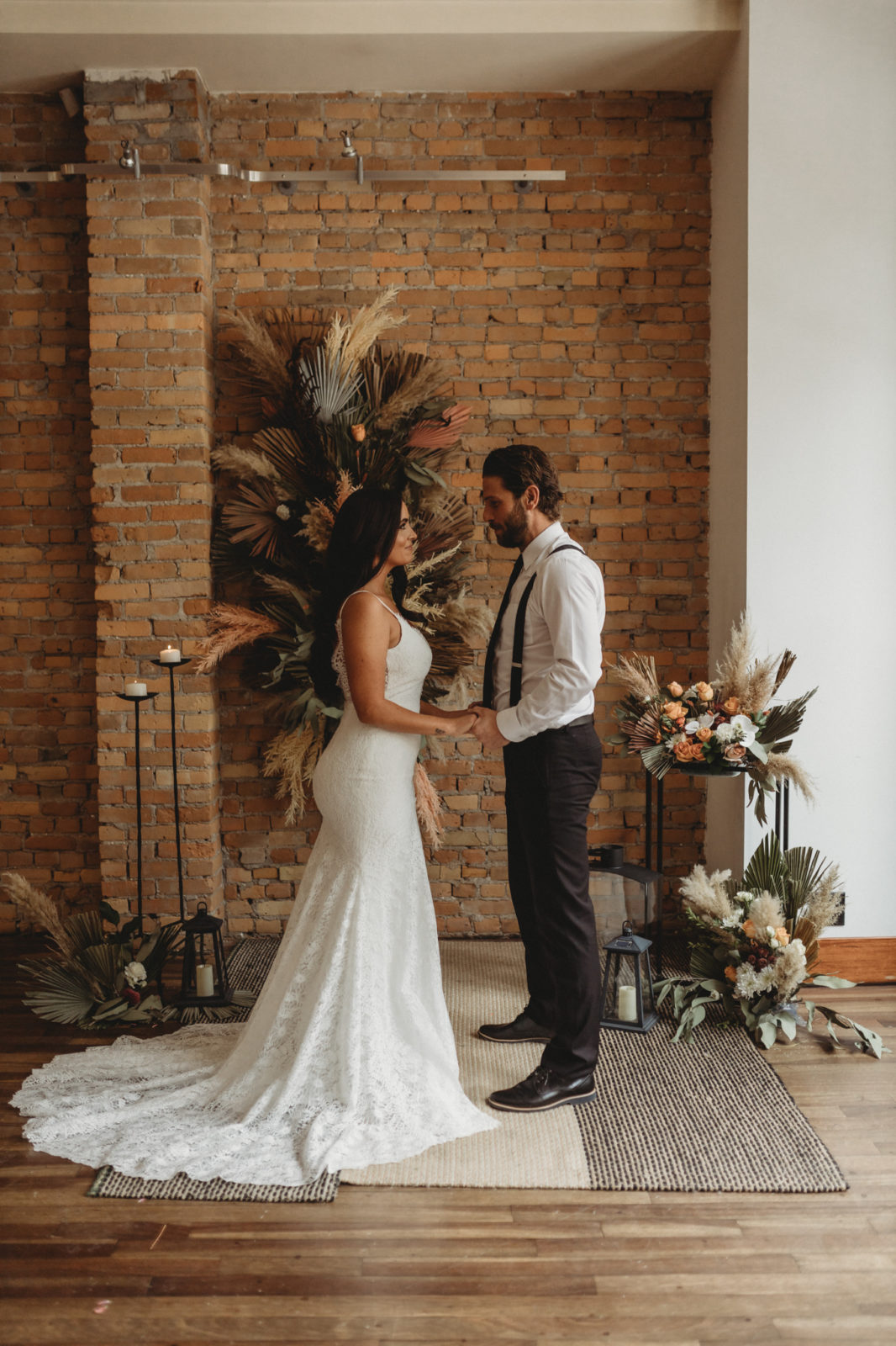 Modern bride and groom holding hands in front of a dried floral installation as wedding ceremony decor inspiration at The Garret