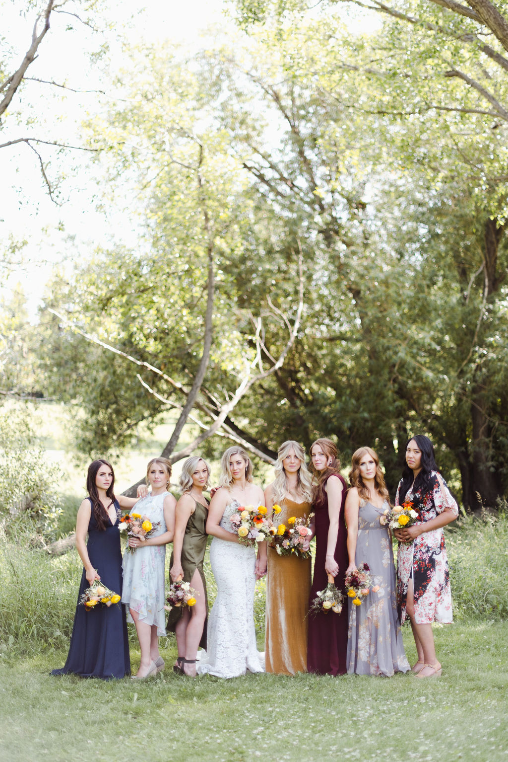 Bridesmaids in mismatching modern gowns pose with the bride on her wedding day