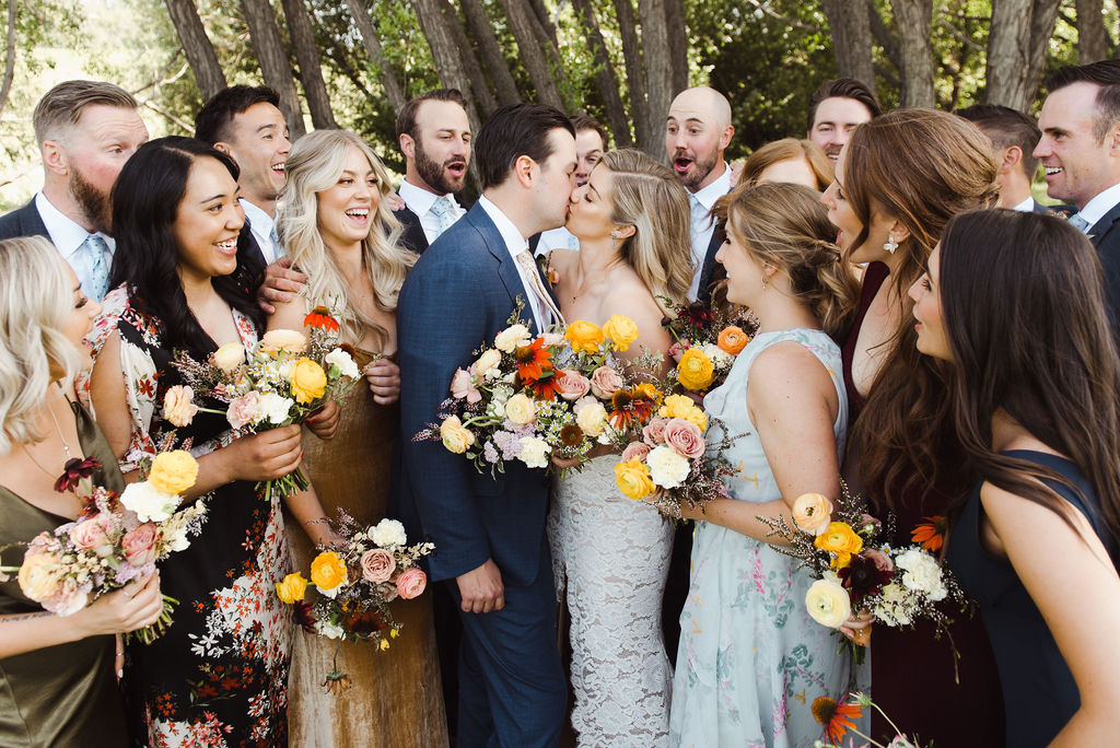 Bride and groom with whimsical wedding flowers share a kiss surrounded by their groomsmen and bridesmaids
