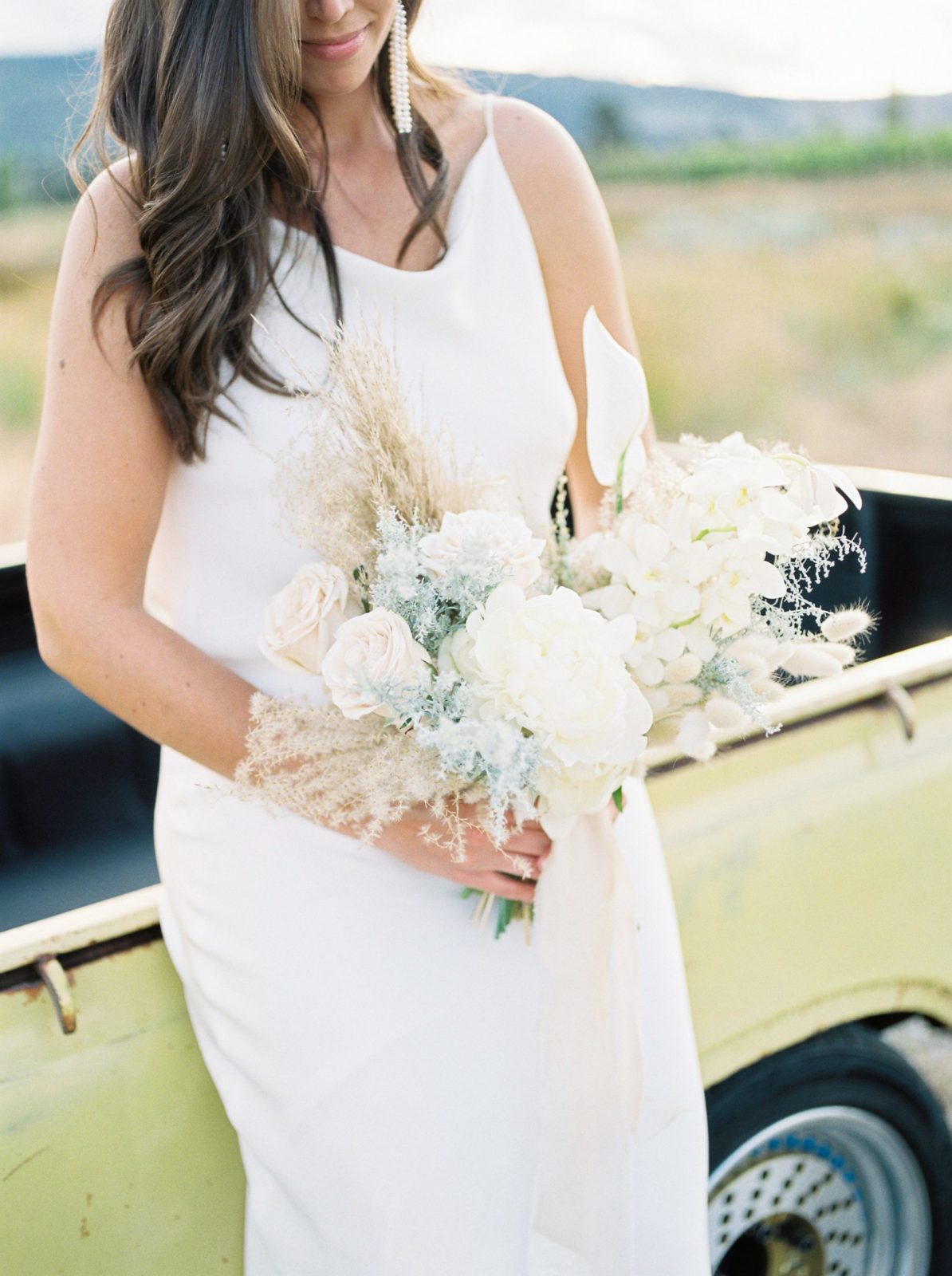 Modern bride holds her boho bouquet while leaning against a pale yellow pickup truck