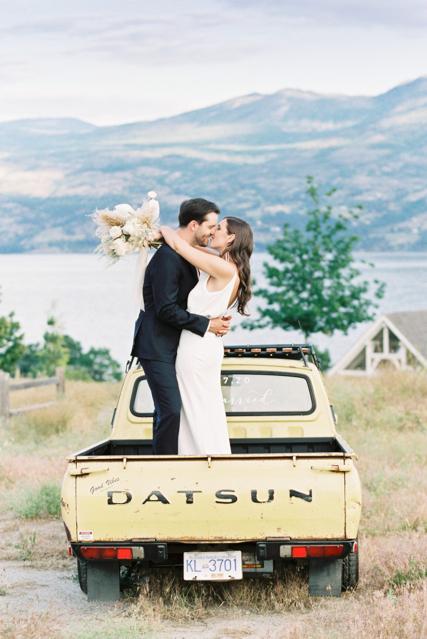 Modern bride and groom share a kiss while standing in the back of their pale yellow pickup truck with the Okanagan mountains in the background