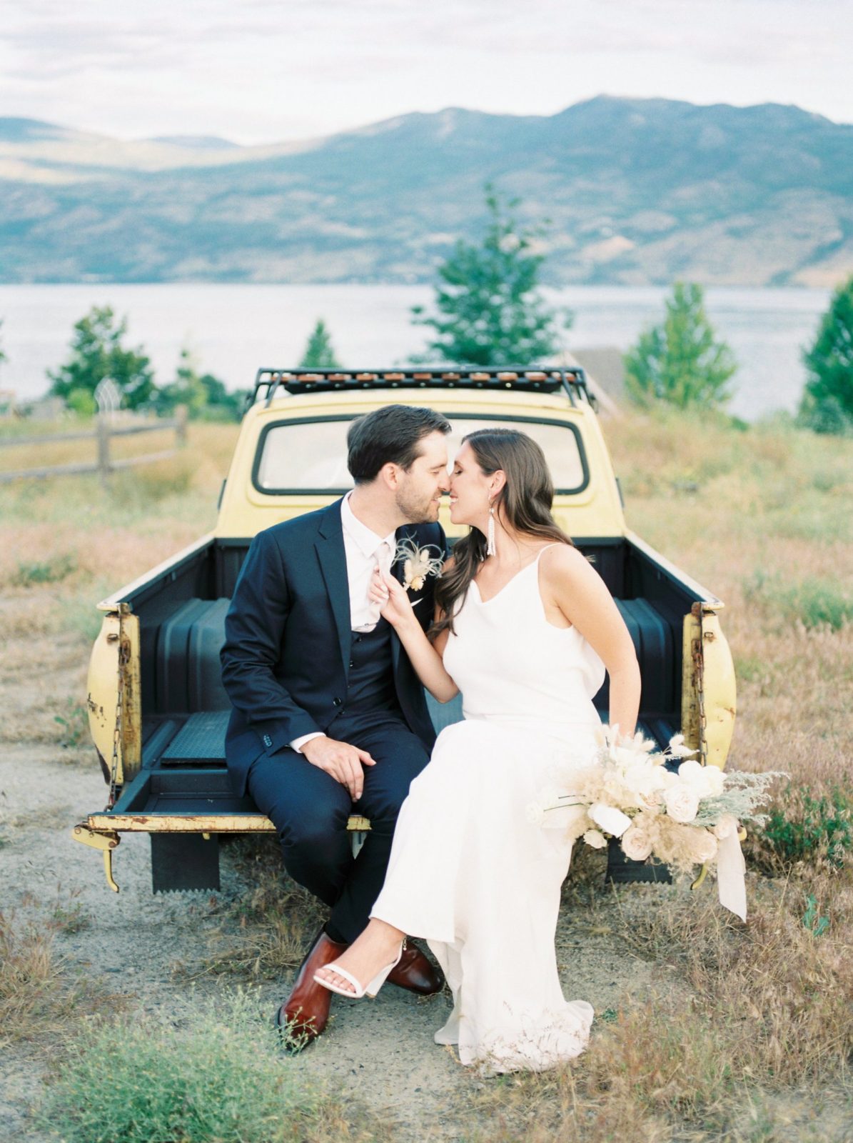 Modern bride and groom pose on the tailgate of a pale yellow truck with a British Columbia mountain range in the background