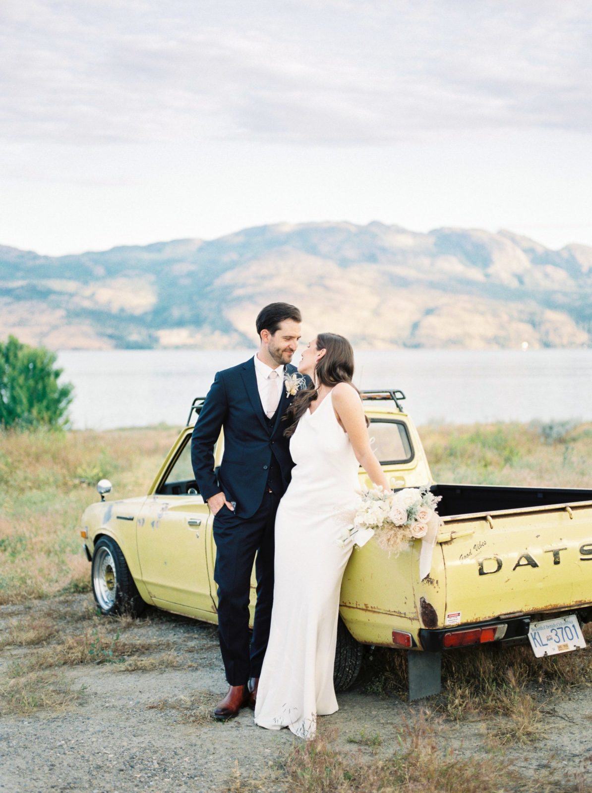 Modern bride and groom pose beside their pale yellow pickup truck with the Okanagan mountains in the background