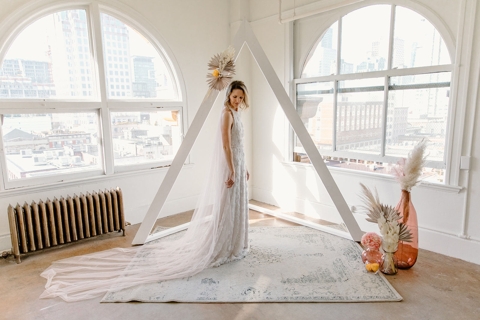 Bride poses in front of a modern ceremony design with a white triangle arch and boho dried florals