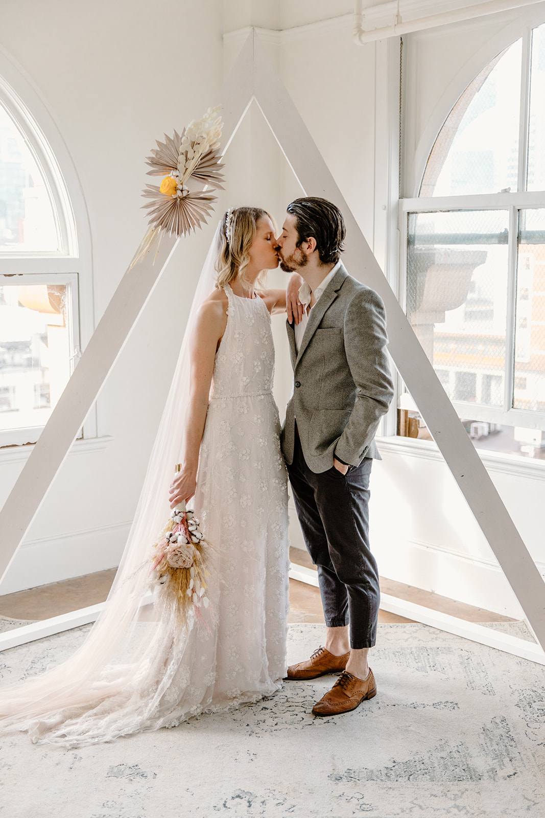 Bride and groom pose under a white triangle arch with dried boho flowers
