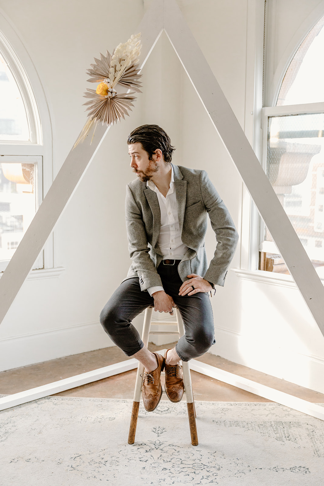 Modern groom poses under a white triangle arch with boho florals