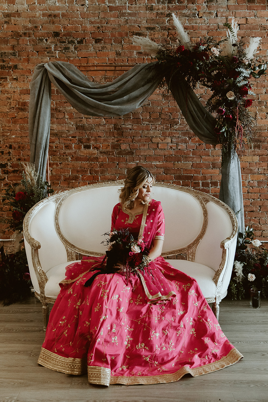 Bride in a bright pink lehenga sits posed on an ornate white loveseat in front of styled ceremony decor at Venue 308 in Calgary Alberta