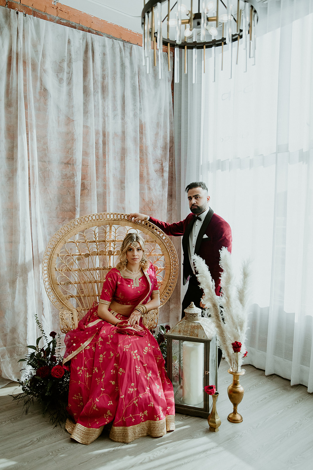 Multicultural bride and groom pose amongst wedding decor featuring wild pampas grass, antique gold details, and a rattan chair for their Indian meets Moroccan styling wedding