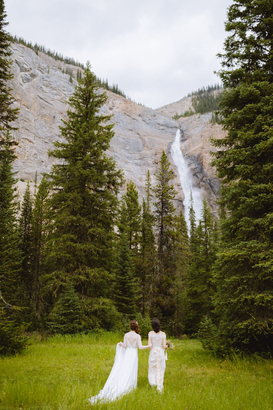 Newly weds in romantic flowing gowns walk towards Takakkaw Falls for their Elopement in Yoho National Park