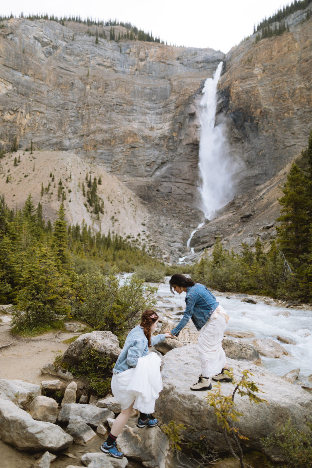 Brides in matching jean jackets help each other climb over rocks with Takakkaw Falls in the background for their Elopement in Yoho National Park