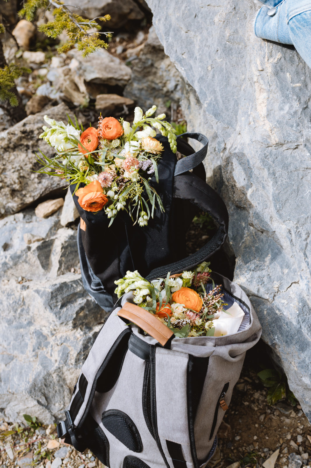 Bouquets designed by Golden Floral Co. featuring bright orange ranunculus sit on top of hiking backpacks against rocks