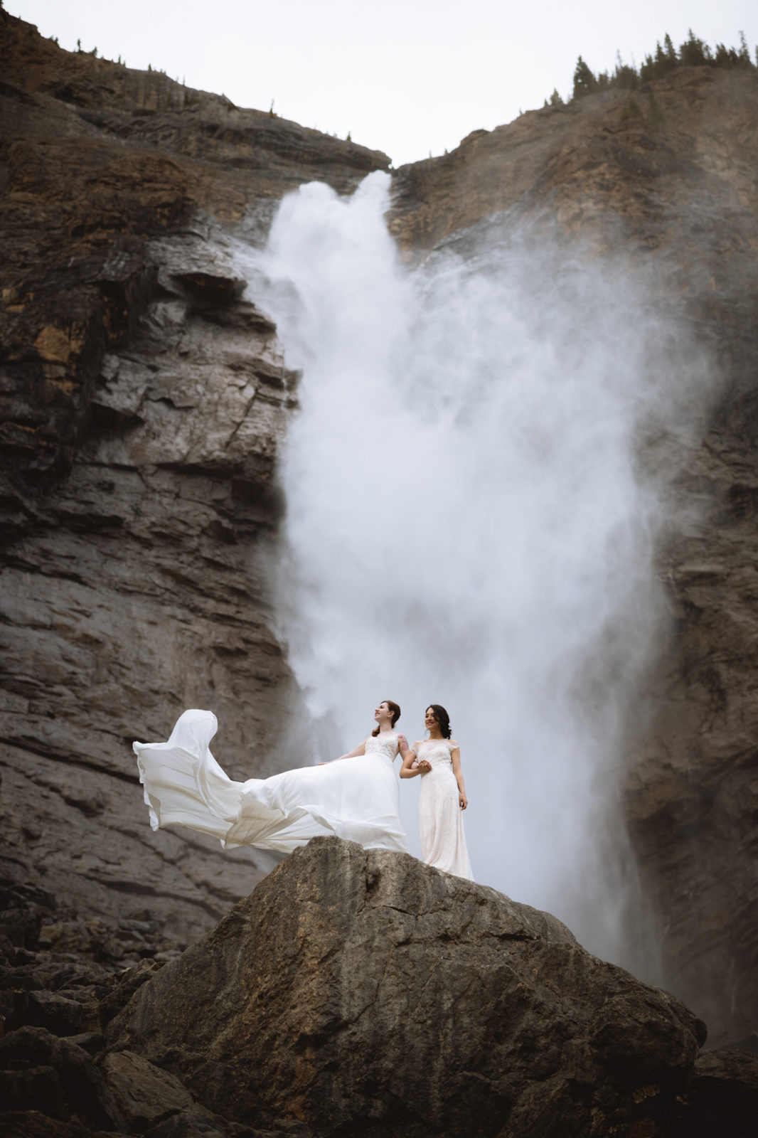 Two brides with romantic gowns billowing, stand in front of a waterfall for their Elopement in Yoho National Park