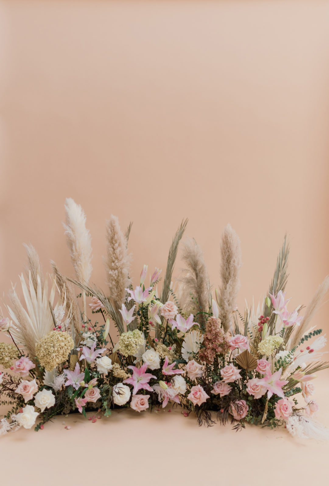 A mix of pink and white blooms, dried grasses, pampas grass and greenery against a peach backdrop