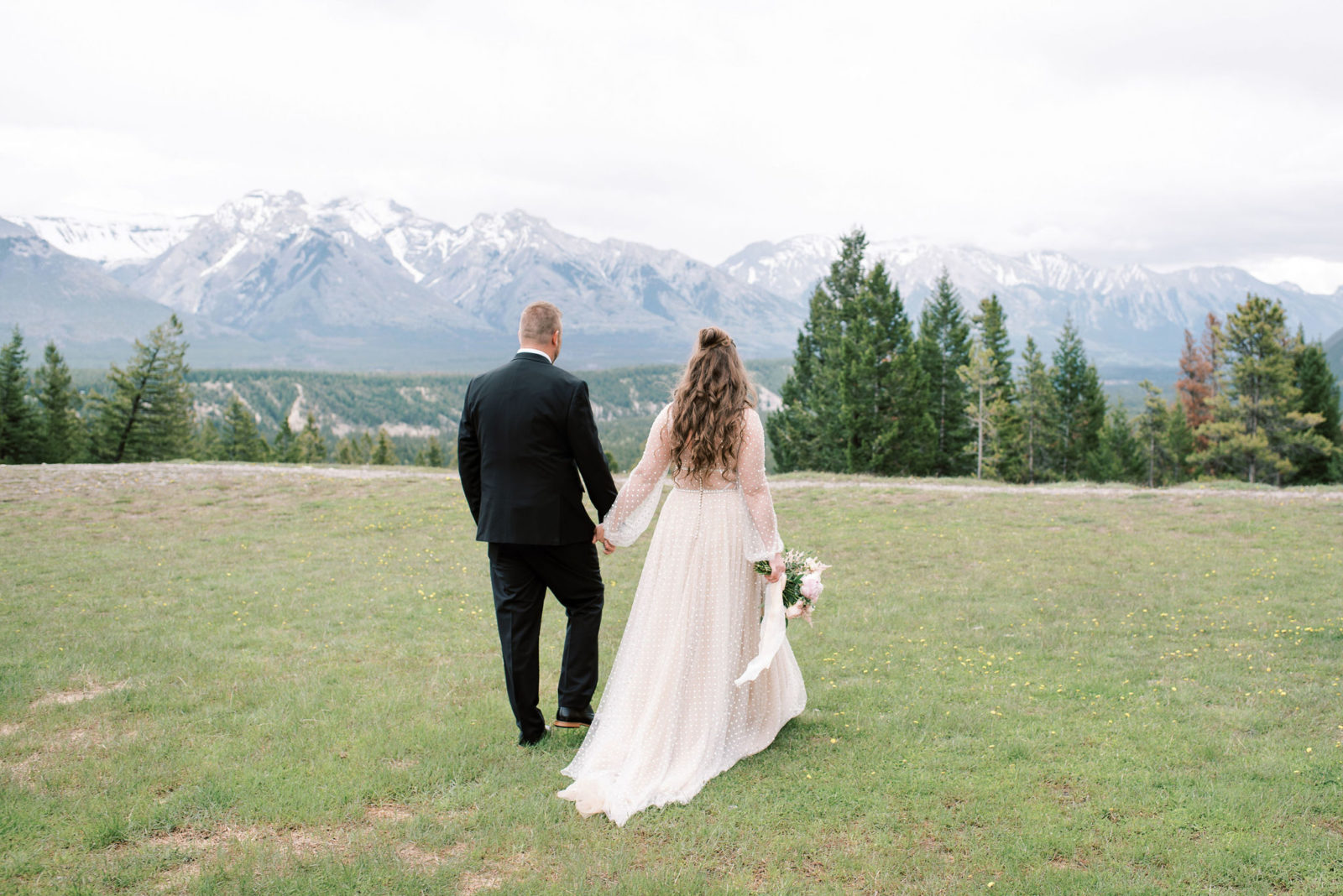 Newly eloped couple in romantic attire walk along the grass at Tunnel Mountain with the Banff National Park mountains in the background