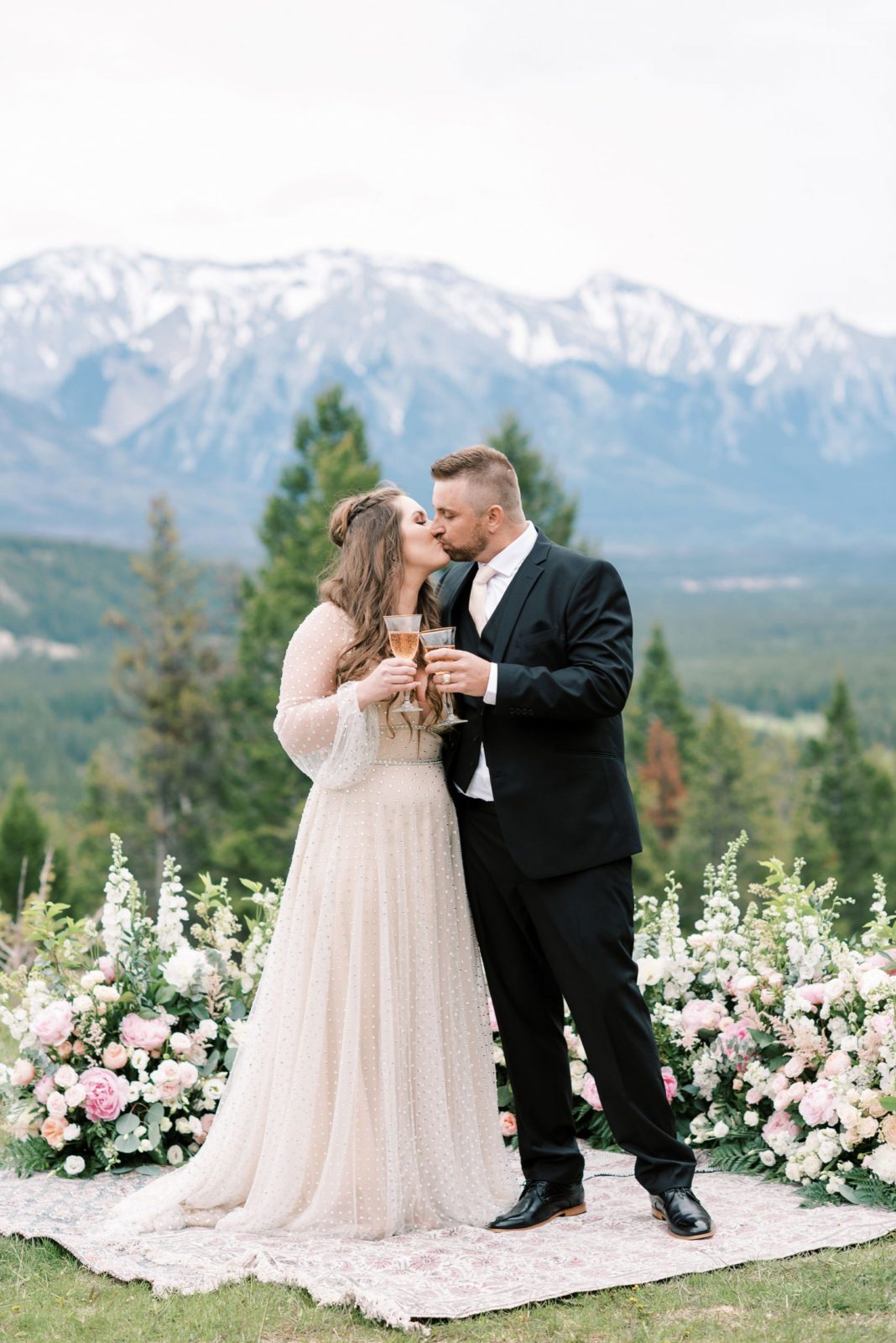 Bride and groom pose with champagne in front of a grounded floral arch created by Flowers by Janie for their mountainside elopement