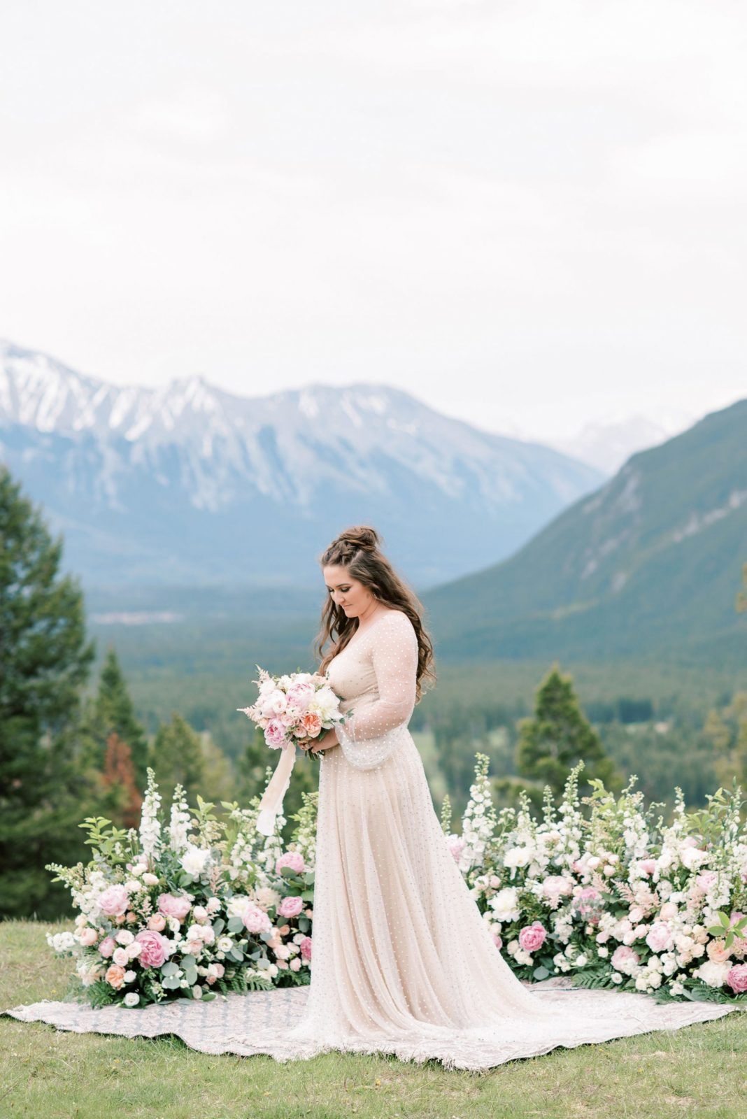 Bride in an oatmeal and blush organza gown by Willowby by Watters holds her blush and pink bouquet at Tunnel Mountain Resevoir in front of a grounded floral arch by Flowers by Janie