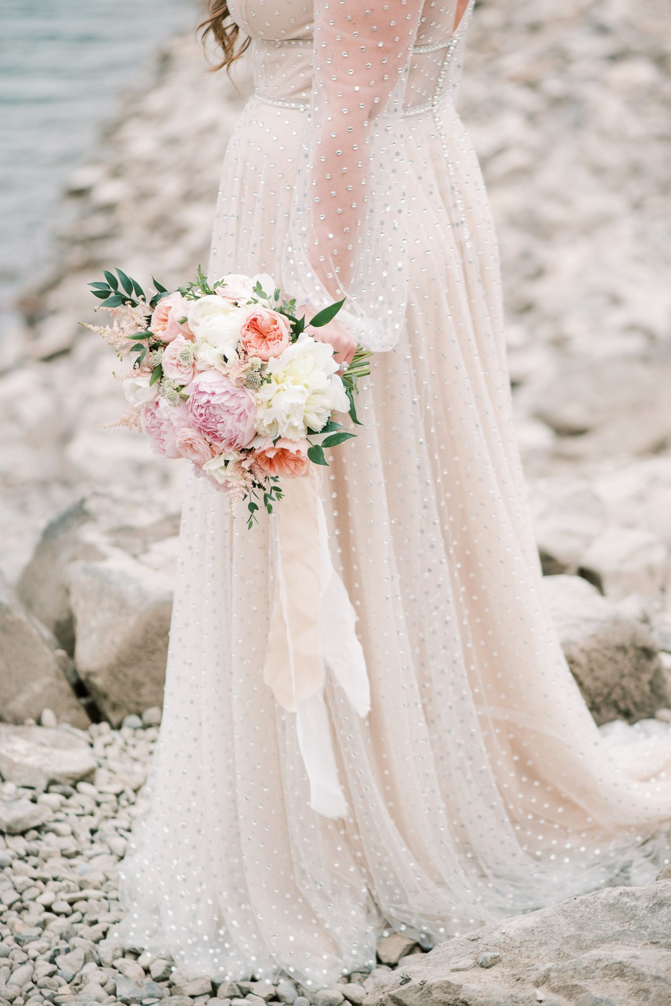 Bride in a blush gown holds her pink and white bridal bouquet on the shores of Lake Minnewanka