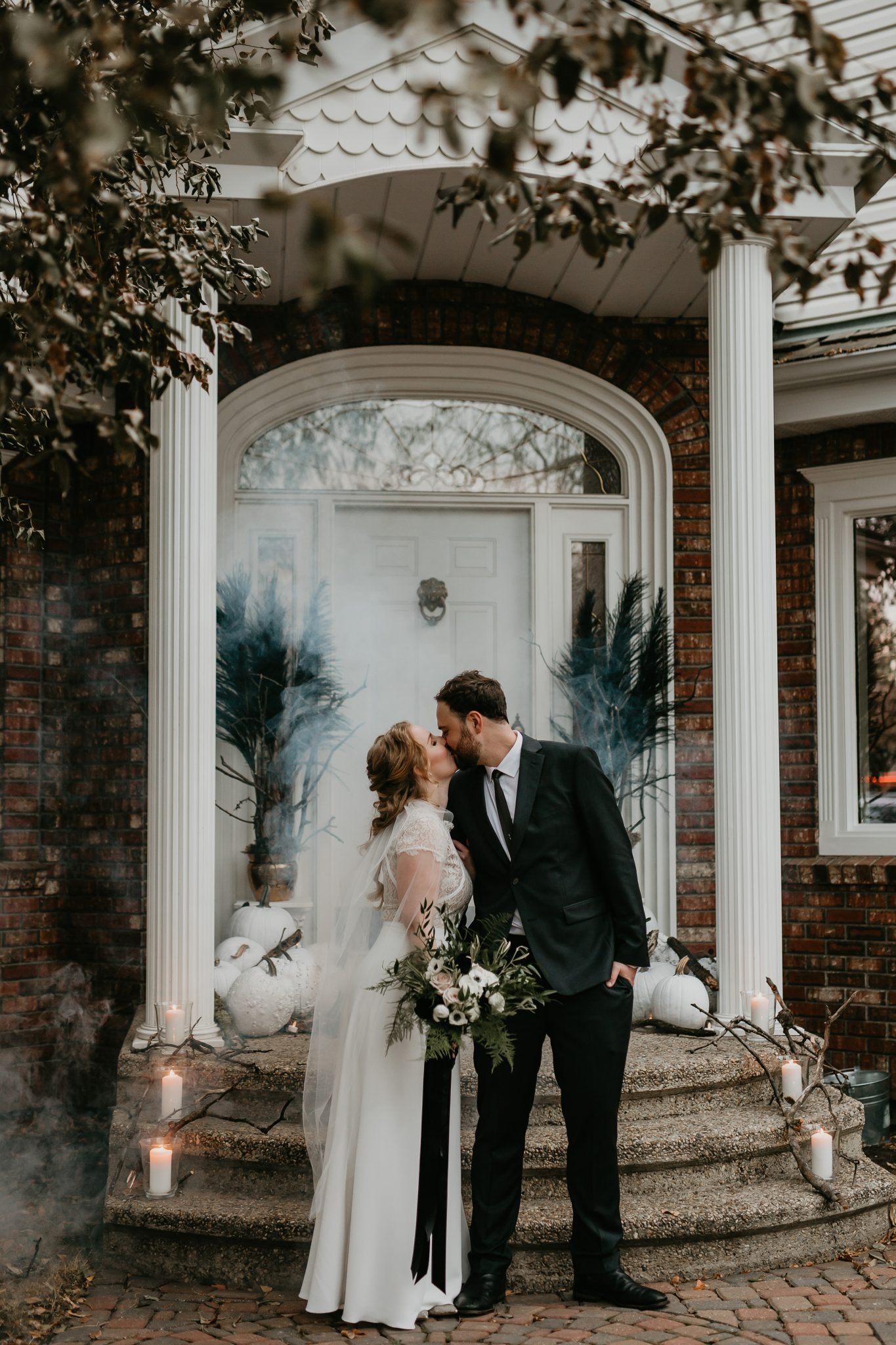 Bride and groom share a kiss in front of their front steps decorated with white pumpkins, a fog machine and candles 
