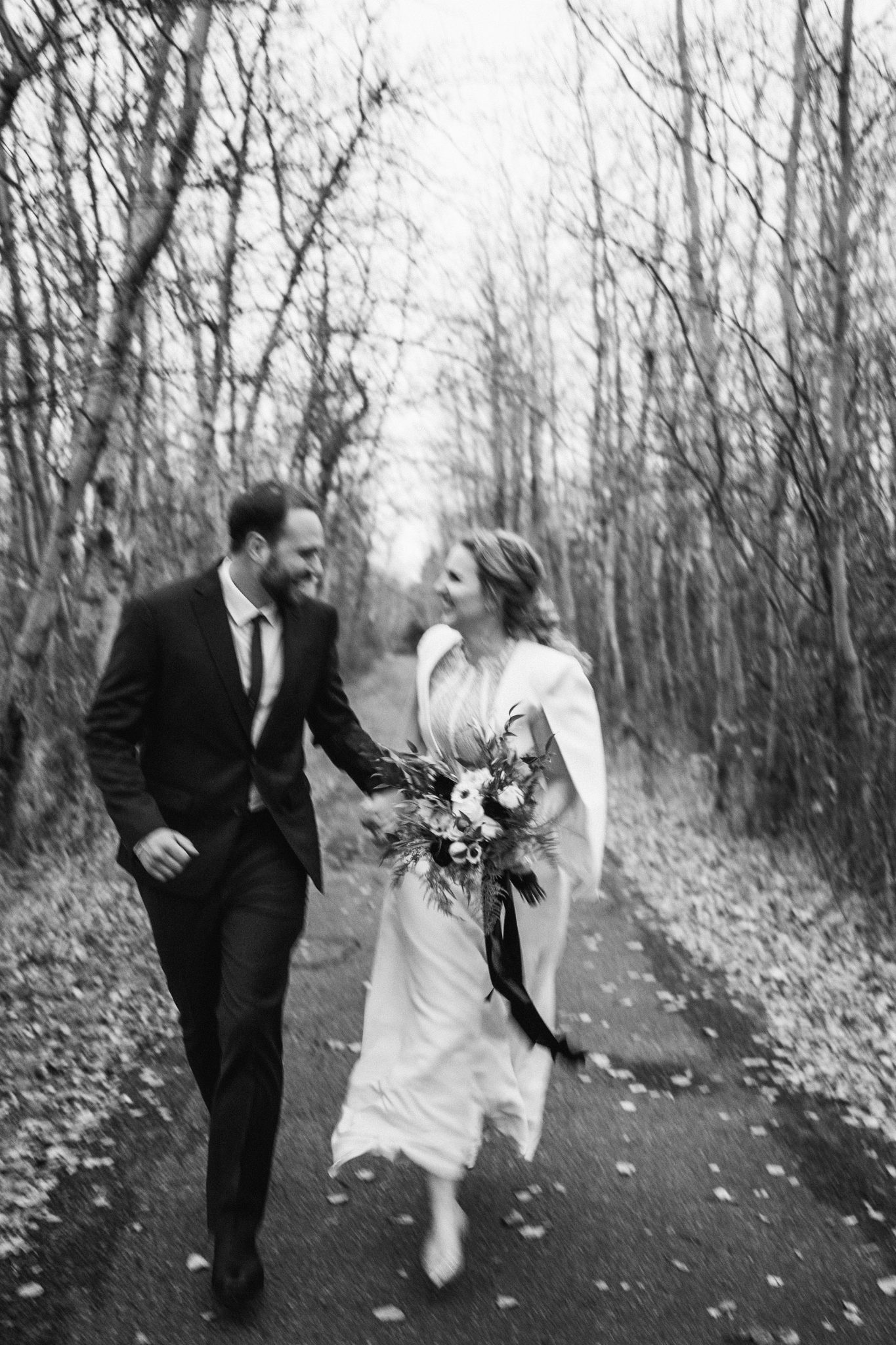 Bride and groom run towards the camera hand in hand down a tree lined path for their October fall wedding
