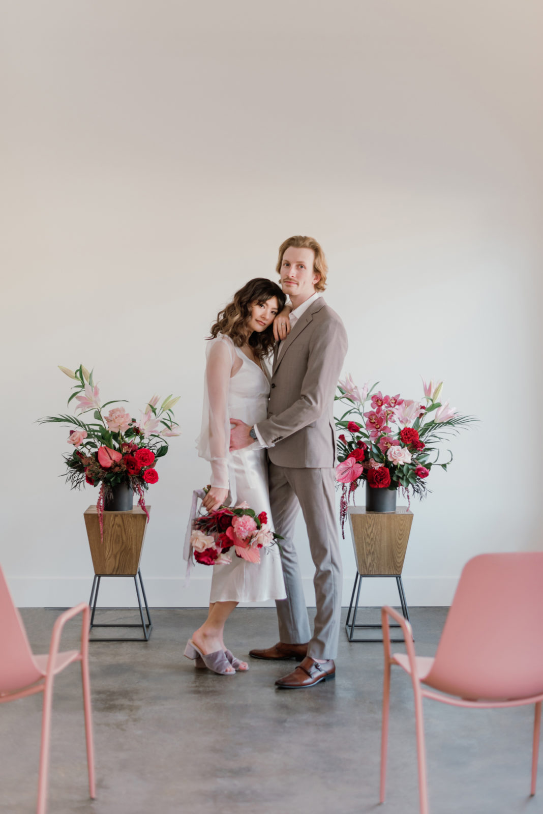 Bride in a short white dress and groom in a grey suit pose in front of berry hued florals for their modern microwedding inspiration ceremony