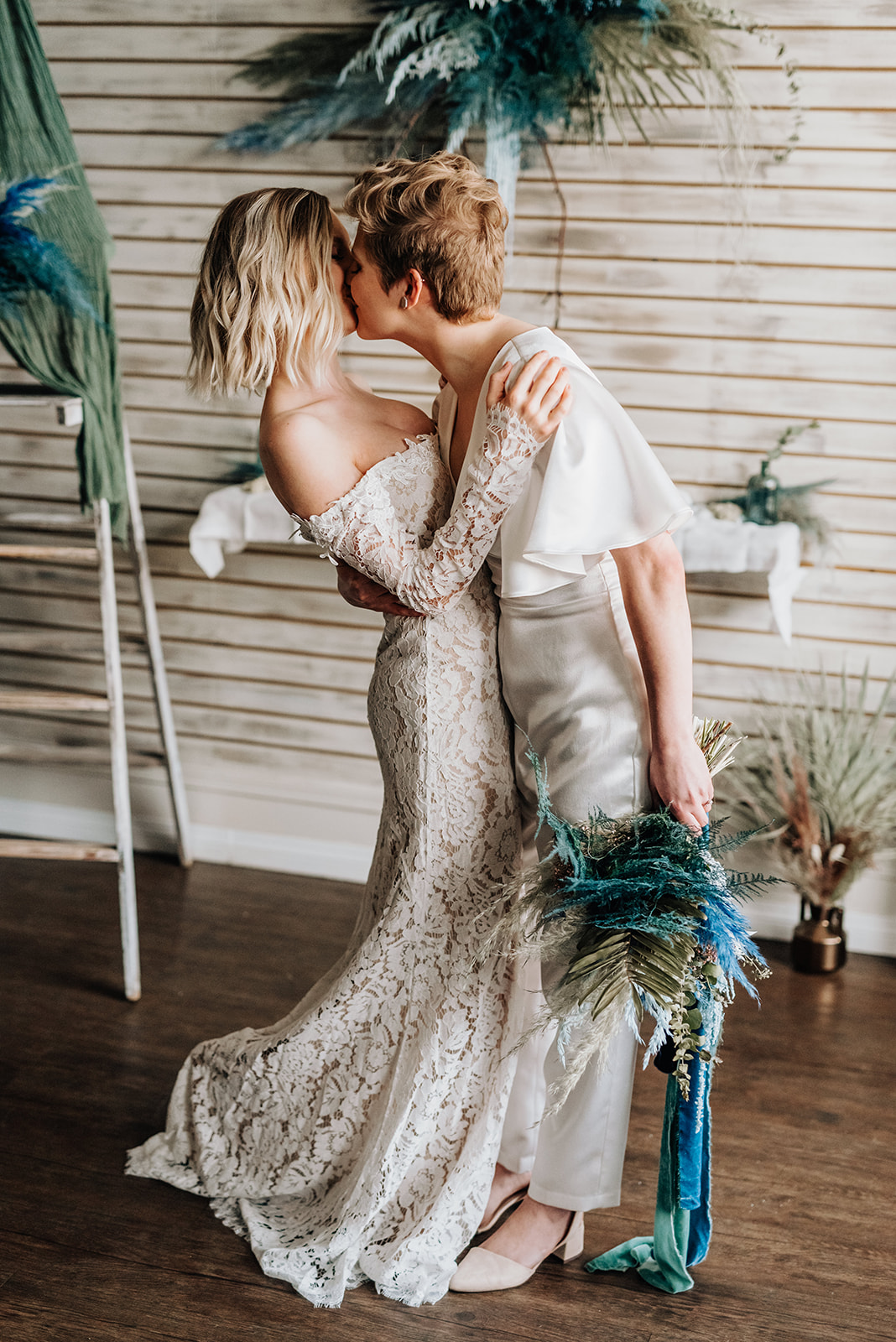 Brides share a kiss in front of their bohemian wedding decor featuring a palette of deep blue and dark teal accents