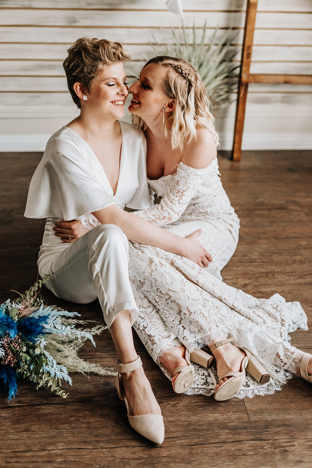 Chic bride in a white jumpsuit sits posed next to her wife in a lace bridal gown