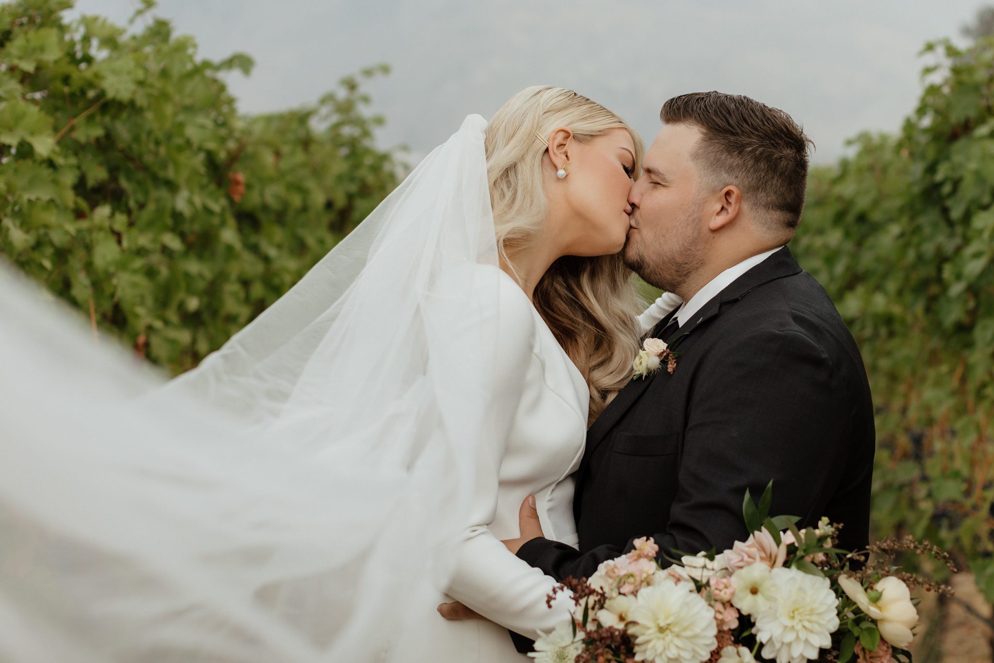 Bride and groom kiss in a vineyard with the bride's long veil flowing freely towards the camera