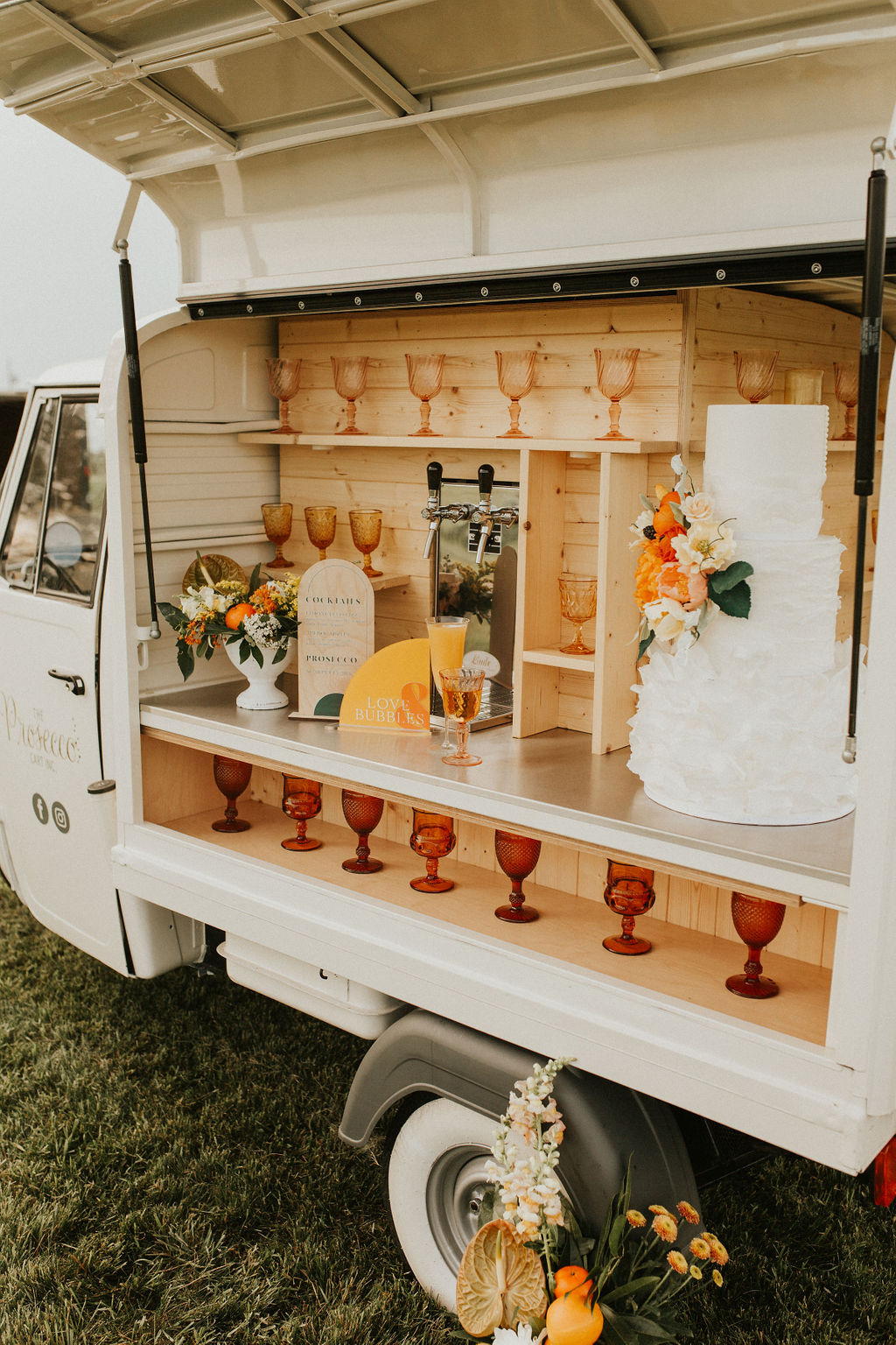 Three tiered ruffled white wedding cake with tangerine floral accents on a mobile prosecco bar cart for wedding decor inspiration