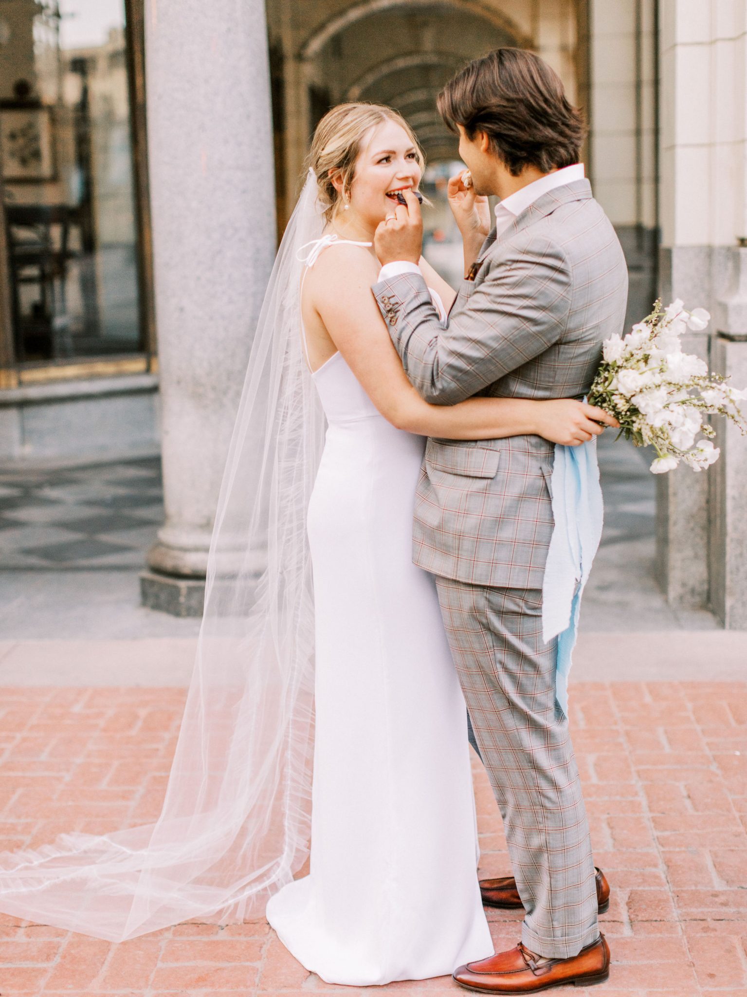 Modern groom in a grey suit enjoys macarons with a bride in a white slip dress