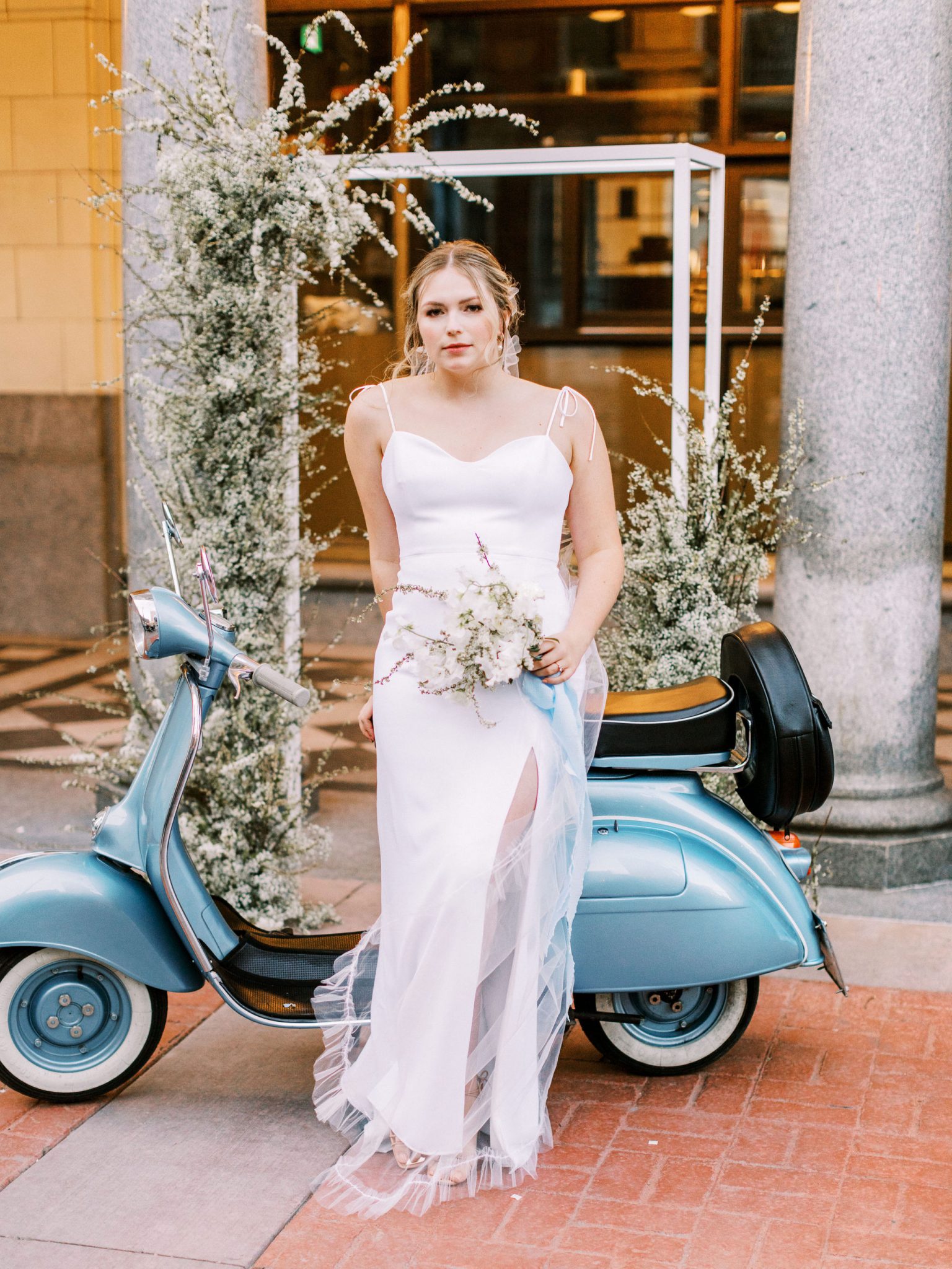 Modern bride in a white slip dress poses in front of a blue vespa with a green and white floral installation behind her for this modern downtown elopement inspiration