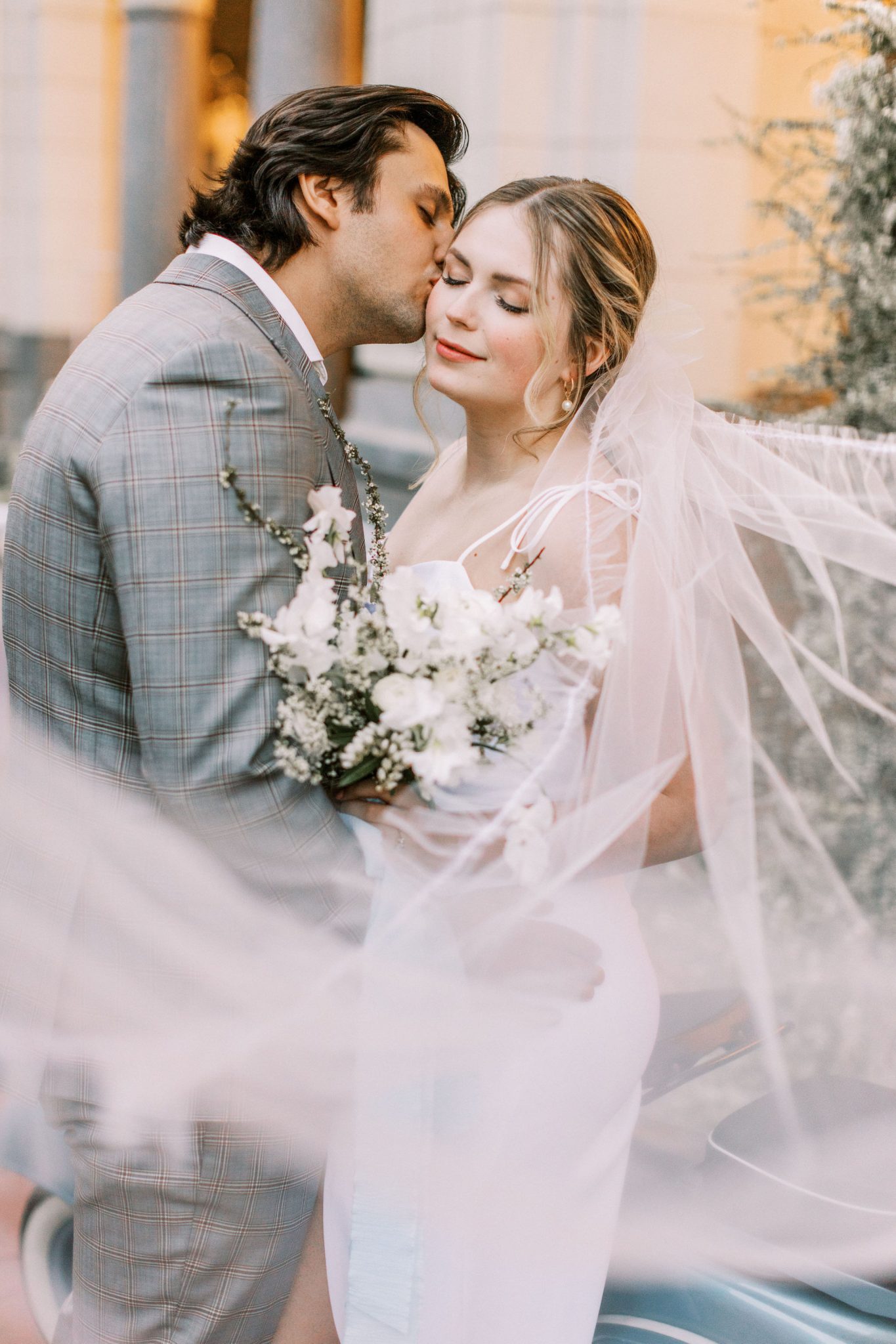 Groom in a modern grey suit kisses his bride on the cheek while her wedding veil flies in the wind