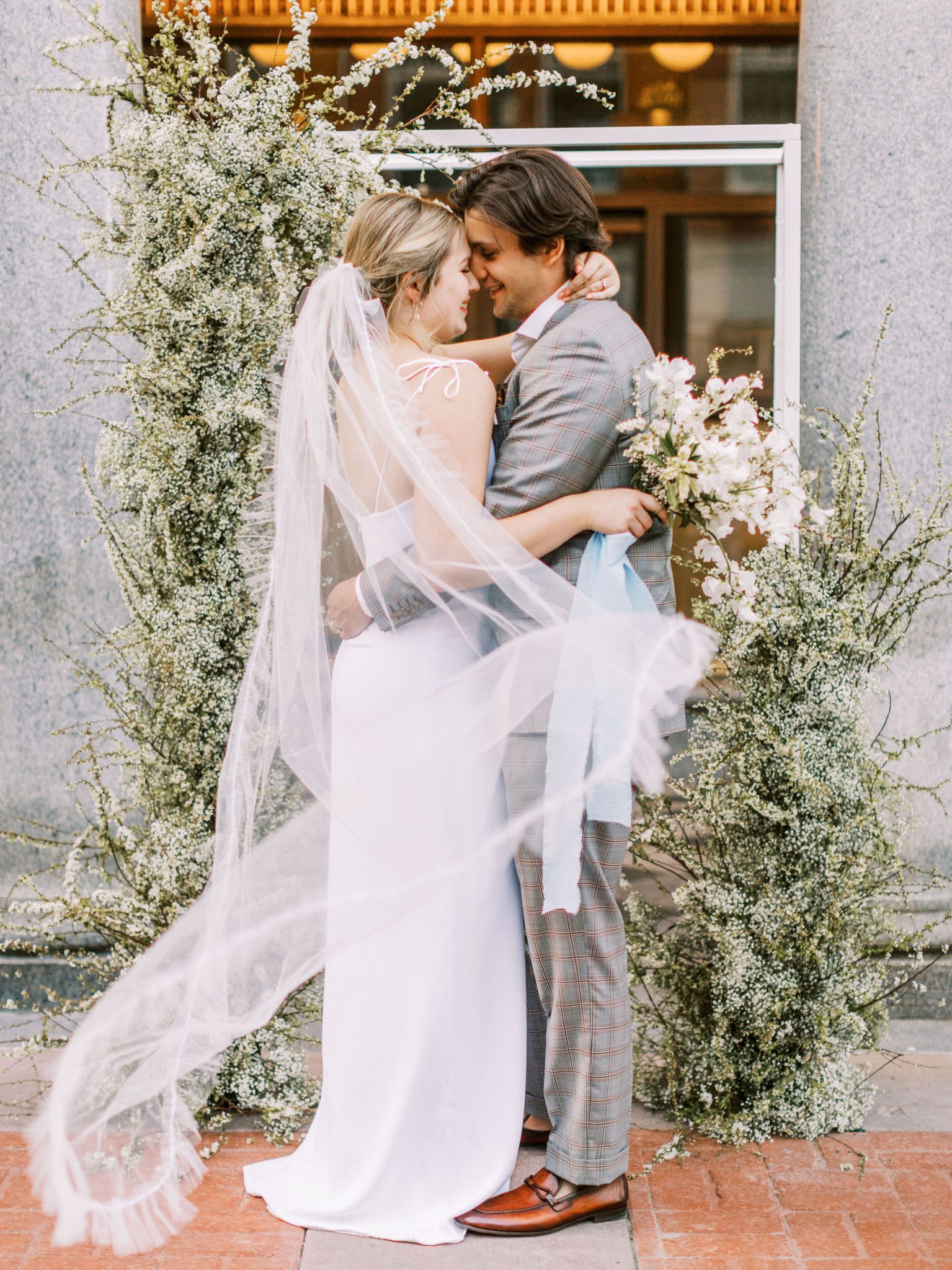 Modern bride in a white slip dress with a flowing veil poses with her grey suit groom in front of a monochromatic floral installation