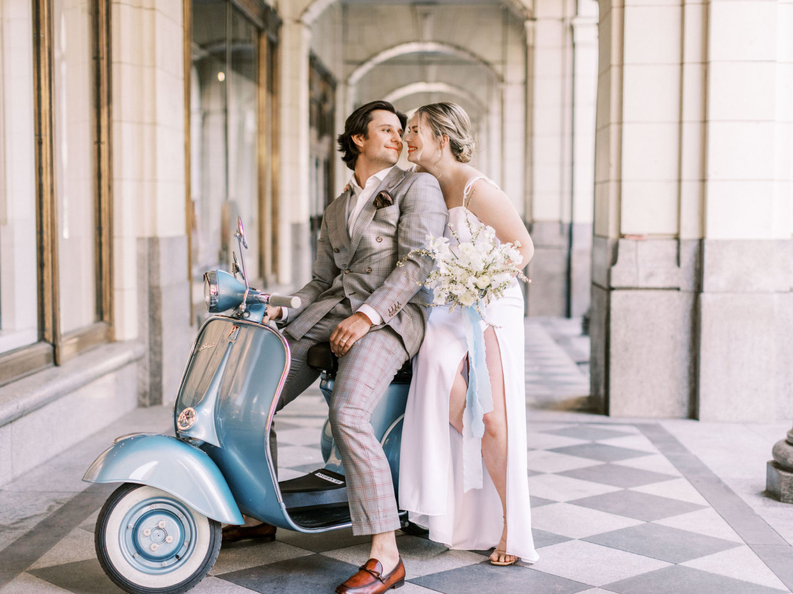 Modern bride in white slip dress and groom in a grey suit pose with a blue vespa for a downtown elopement
