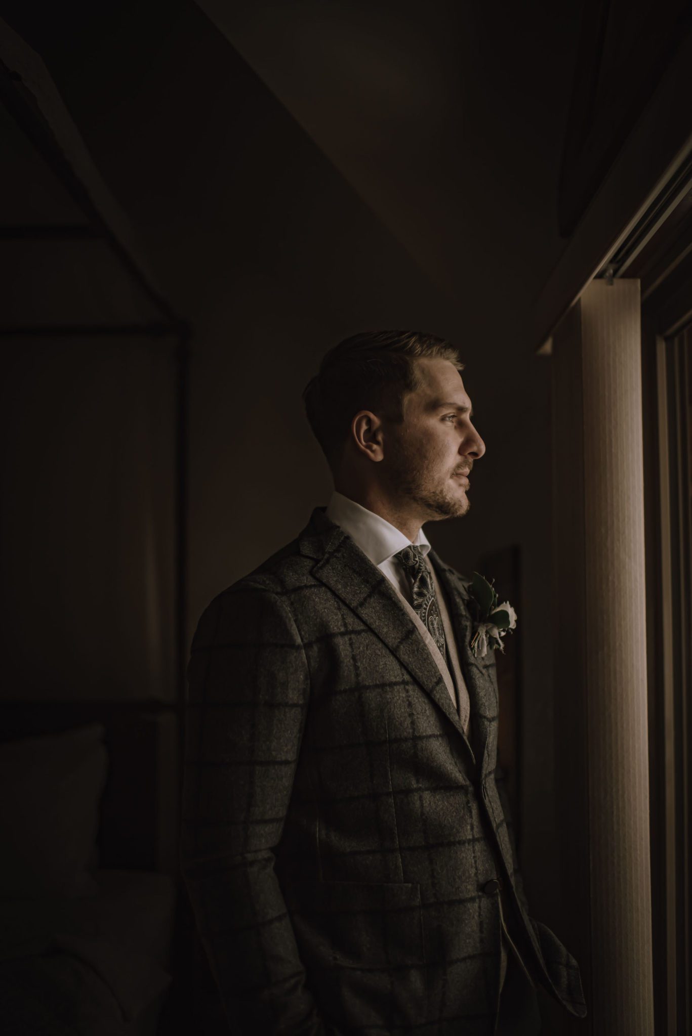 Styled groom in a grey three piece suit looks out the window at A Bear & Bison Canadian Country Inn in Canmore Alberta
