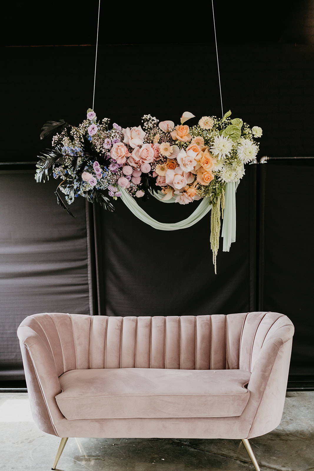 Pink loveseat underneath a colourful floral installation with a black backdrop