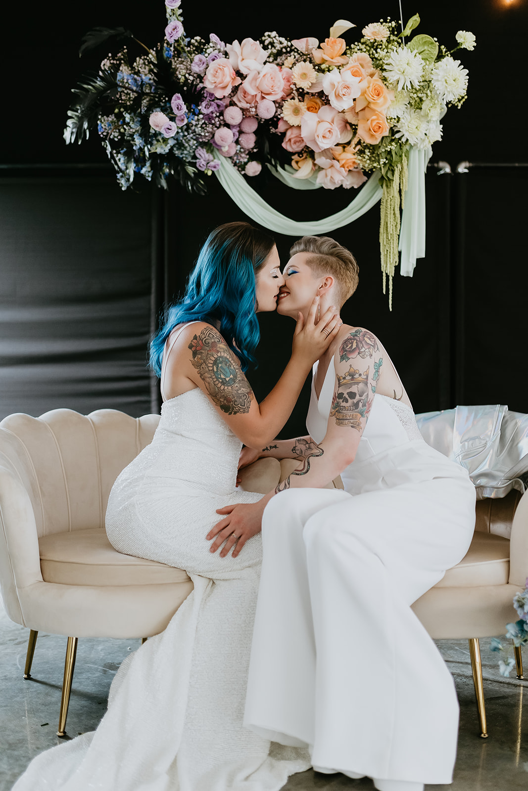 Rocker chic brides kiss under many coloured floral chandelier