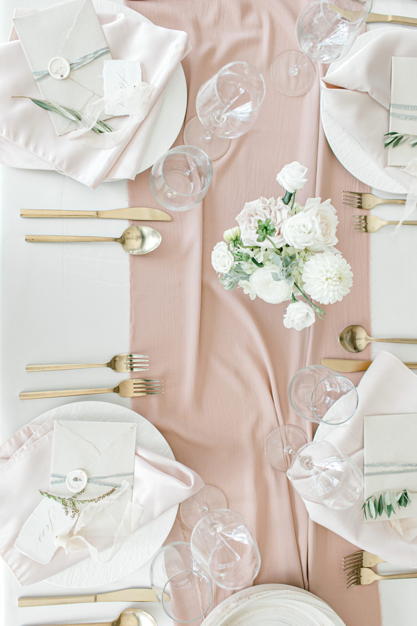 Wedding table photo above showcasing place settings, gold cutlery, blush linens, white and green florals and greenery, altogether a classic yet modern wedding table design