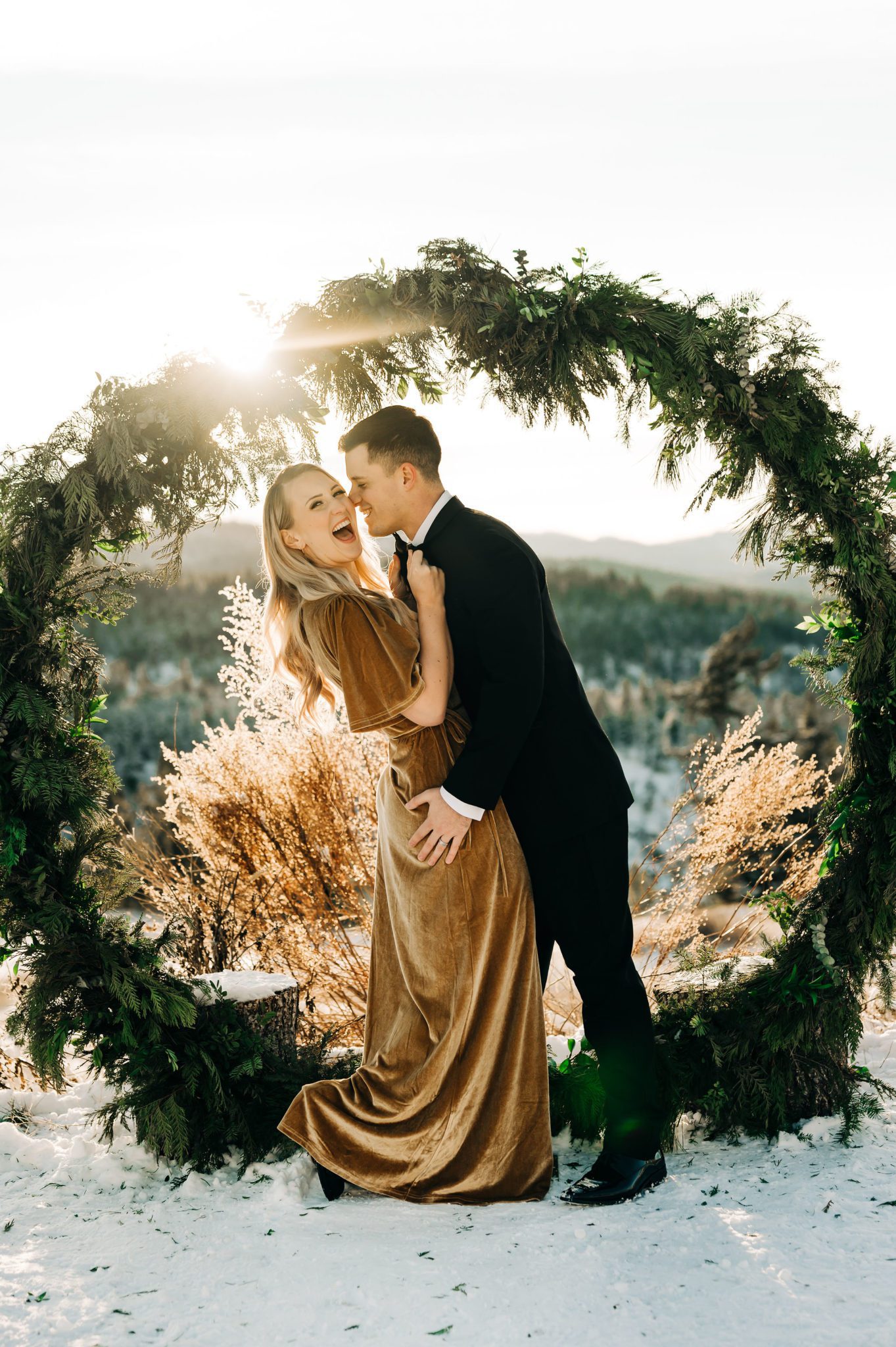 Couple poses under pine archway at golden hour elopement, wedding attire inspiration in mustard velvet dress