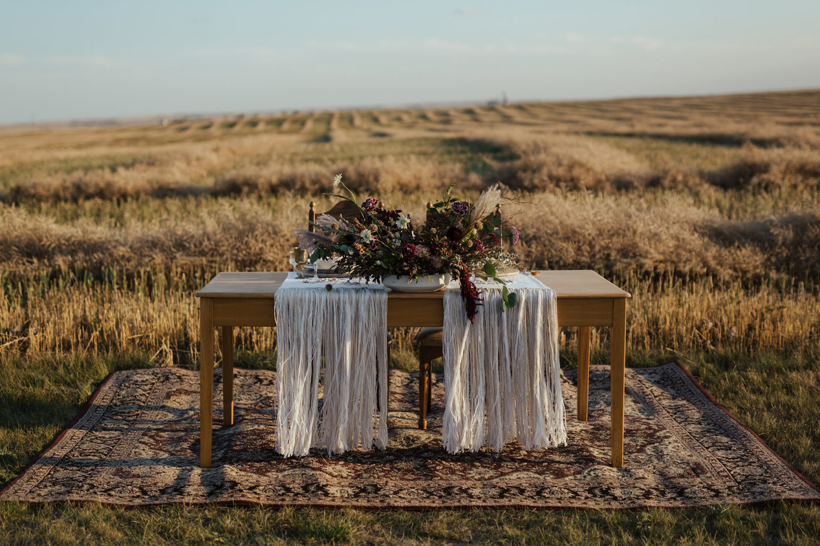 Autumn-inspired tablescape with rich coloured floral arrangements, placed on top of a wooden sweetheart table for the bride and groom, with cream-coloured drapery and vintage glassware, located in the rural Alberta countryside, fall vow renewal inspiration. 