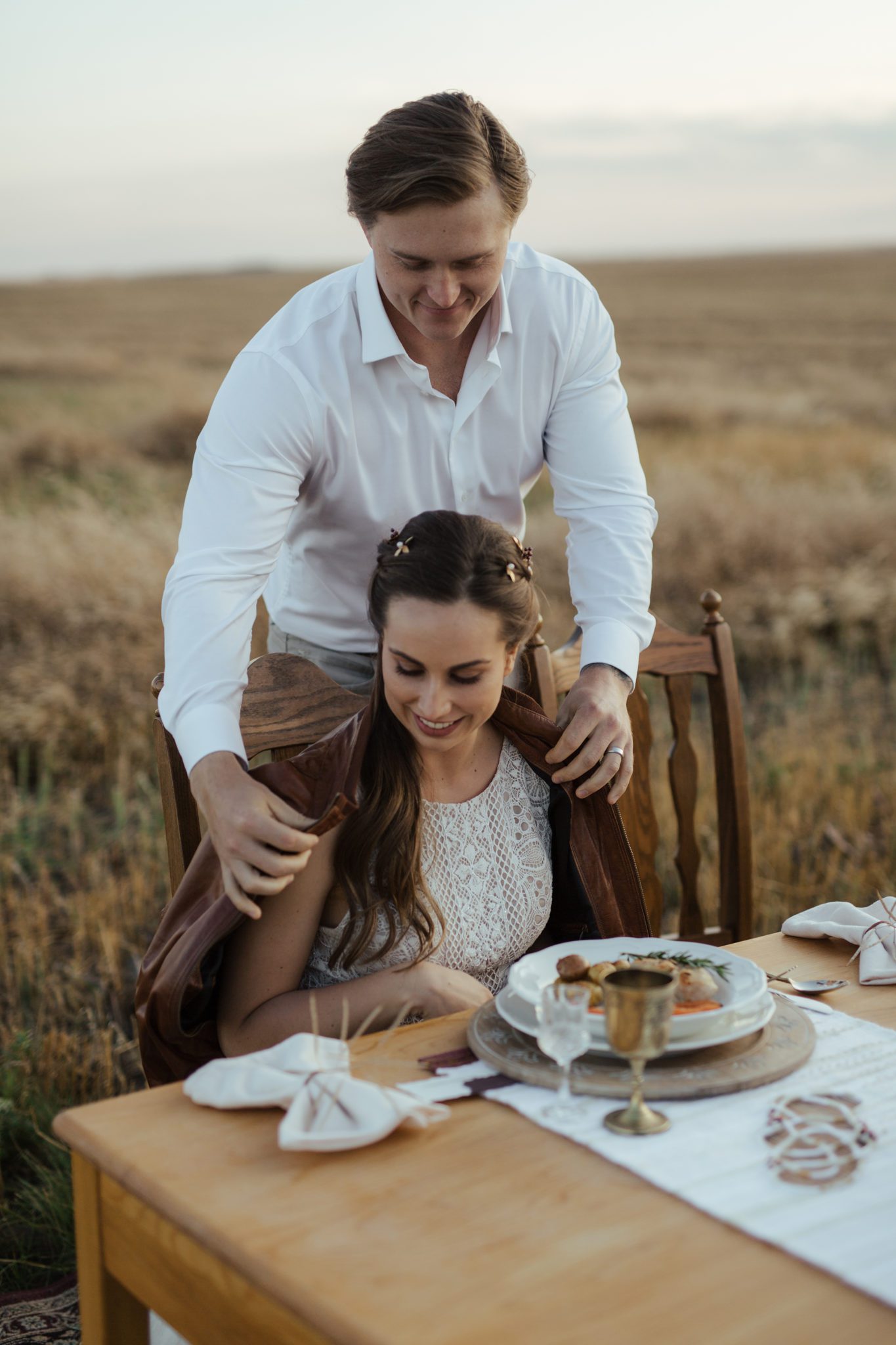 Portrait of groom putting his brown-coloured jacket on his bride while she sits at their autumn-inspired sweetheart table, located in the rural Alberta countryside, fall vow renewal inspiration. 