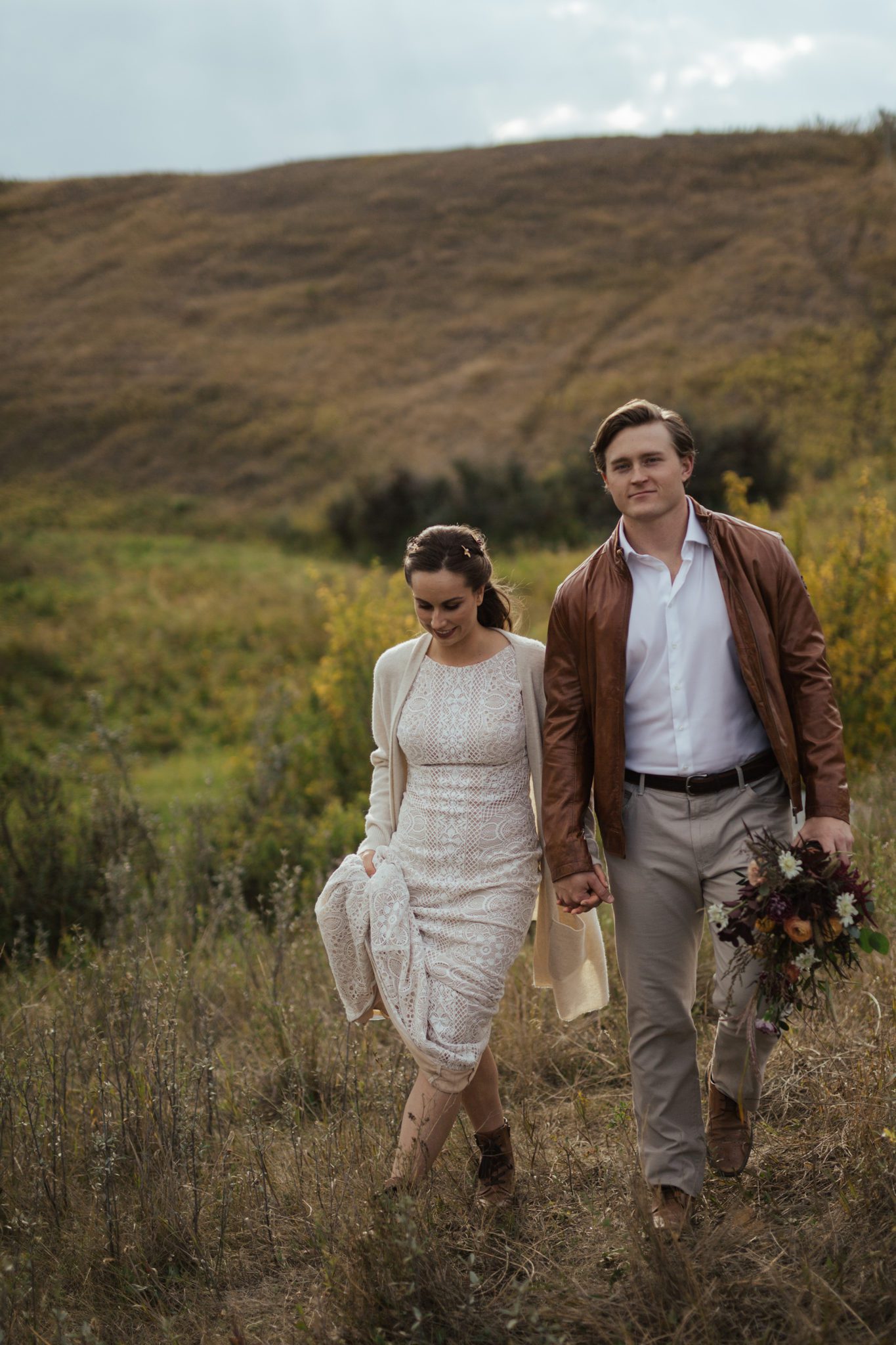 Bride and groom walking in the countryside of rural Alberta, while bride is wearing lace-detailed gown and groom has a brown-coloured jacket, featuring fall-coloured bridal bouquet, fall vow renewal inspiration. 