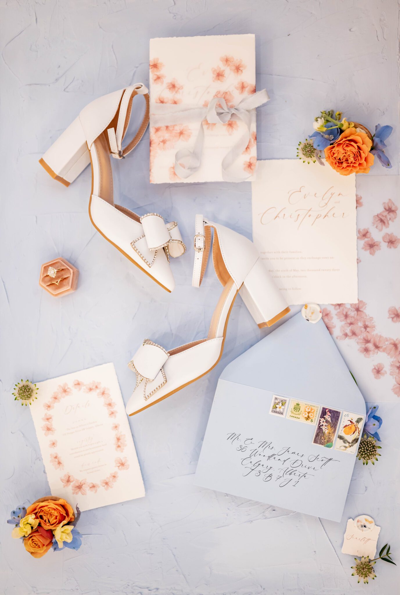 Wedding invitation and place card stationery flat lay featuring calligraphy and delicate flower details wild flower wax seals and a baby blue envelope , feminine pointy-toe bridal shoes with large bow, gold engagement ring and wedding band showcased in light pink velvet ring box