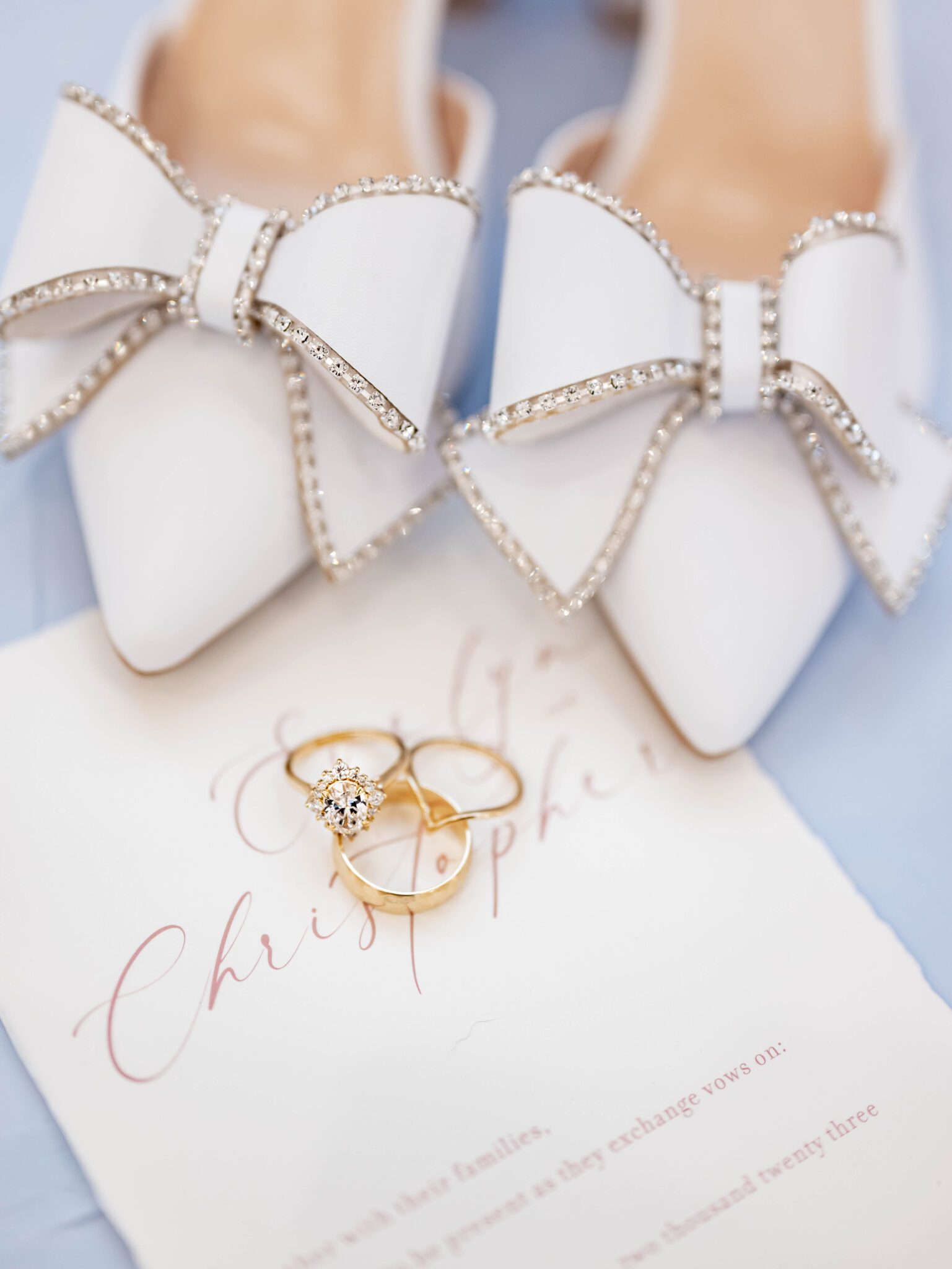 Elegant white pointy-toe bridal heels featuring a large bow adorned with sparkling crystals, wedding invitation flat-lay inspiration with bridal shoes and rings