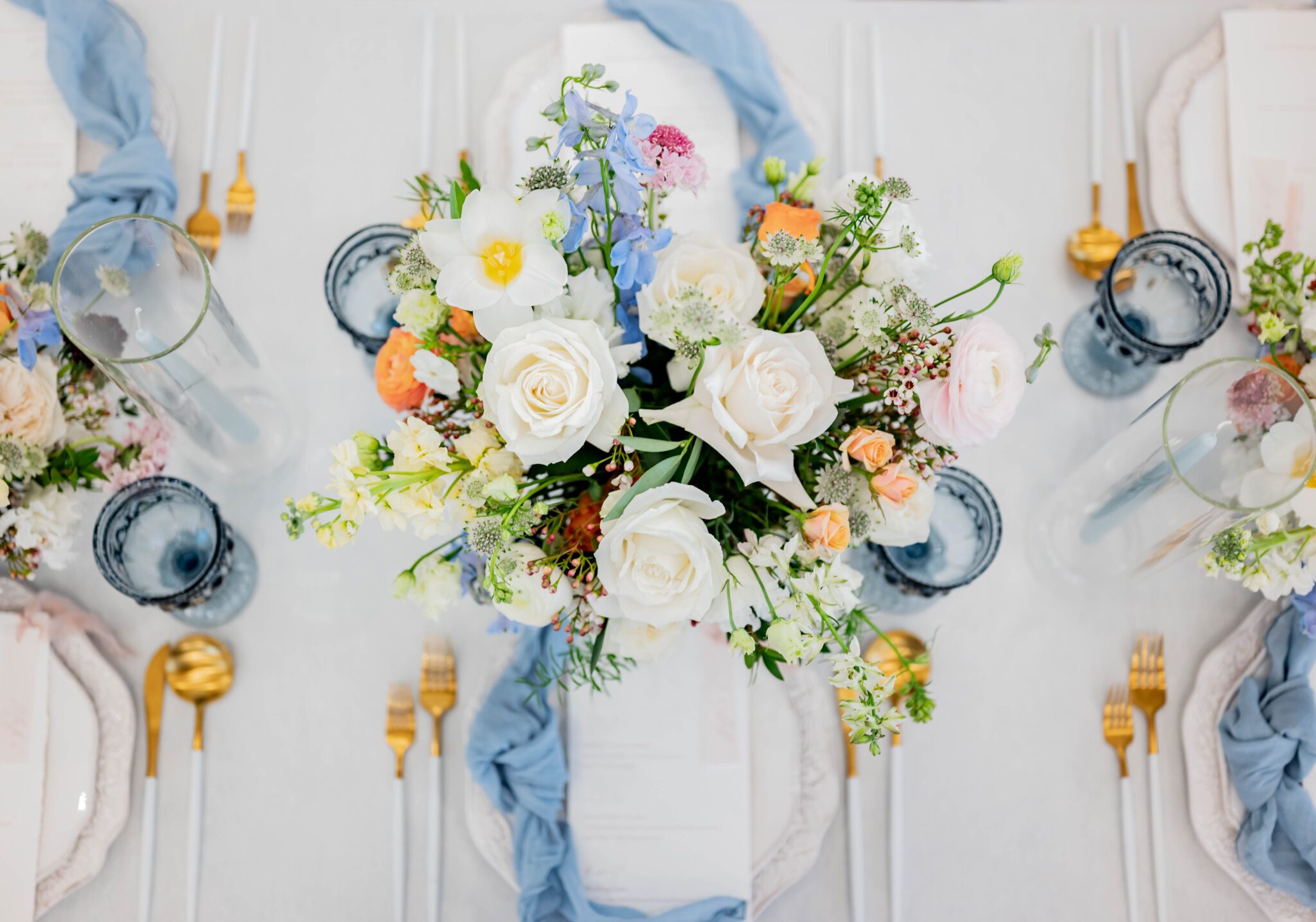 Wildflower wedding inspiration featuring pastel-inspired guest tables with charming floral arrangements, guest table adorned with white and gold cutlery and baby blue napkins delicately tied in a loose knot