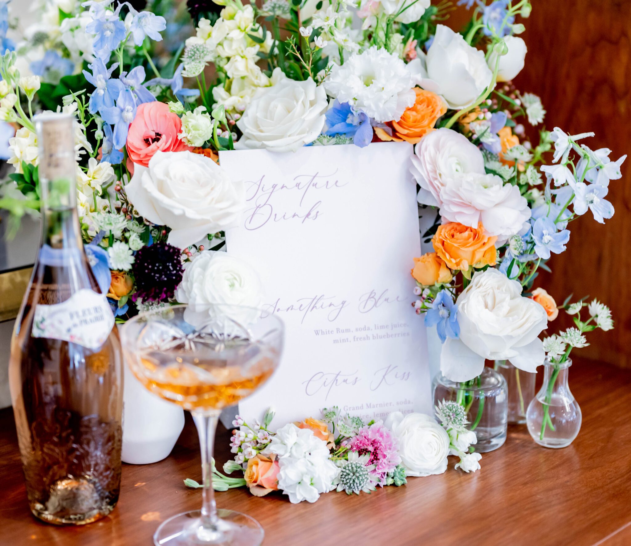 Signature drink menu written in calligraphy on wood bar top, bar menu display with spring wild flowers in delicate tones of blue, orange and green, bubbly rose in crystal coup glass displayed by signature drink menu