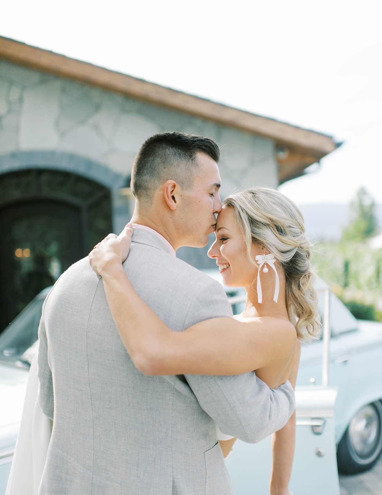 Classic car parked in front of an idyllic summer wedding venue, groom kisses bride's forehead ready to whisk her away in style, loose curls pulled back in a glamorous bridal hairstyle by Kathryn Ramsey Esthetics showcasing white bow earrings