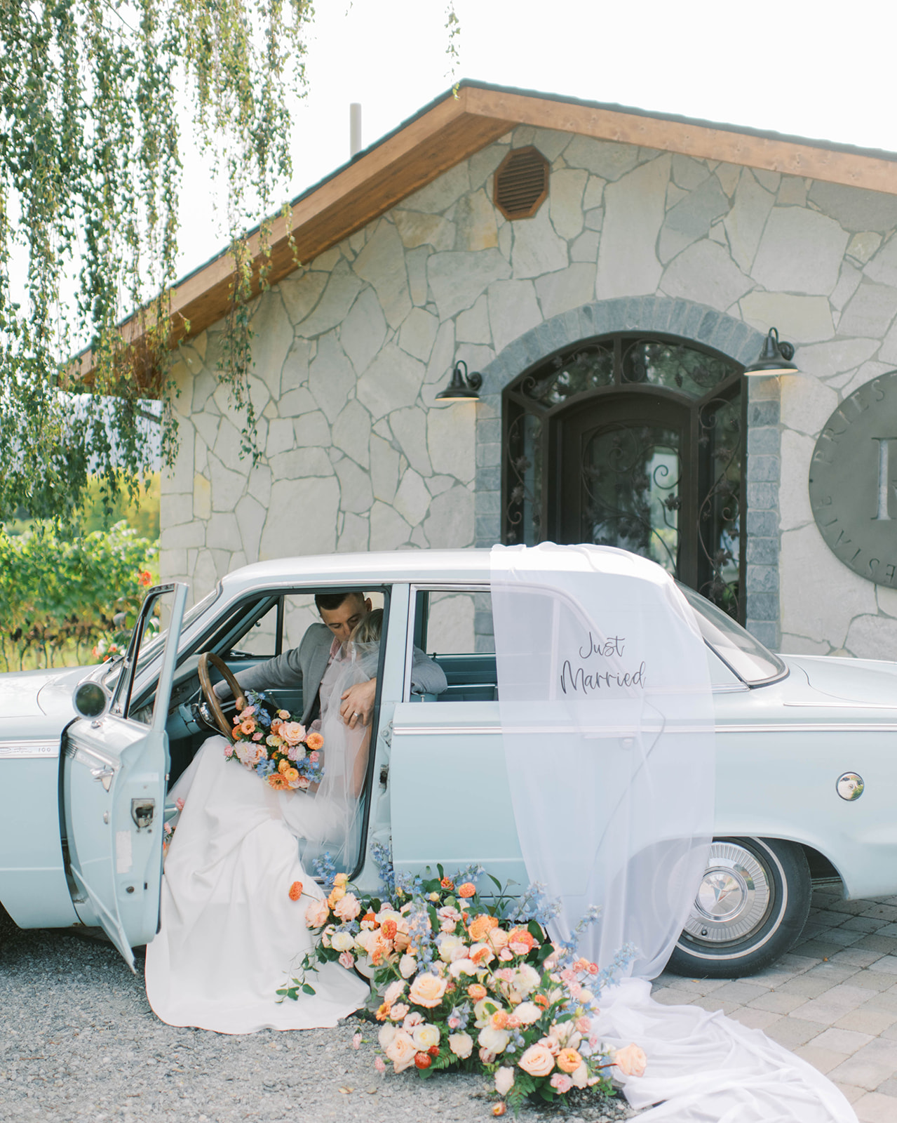 Bride's veil gracefully draped on blue vintage car with just married sign by Aspen Etched, adding a touch of romance to the getaway vehicle, lush floral arrangements adorning a classic car, transforming it into a stunning centrepiece for a vintage-inspired wedding, bride and groom sharing an intimate moment in blue getaway car 