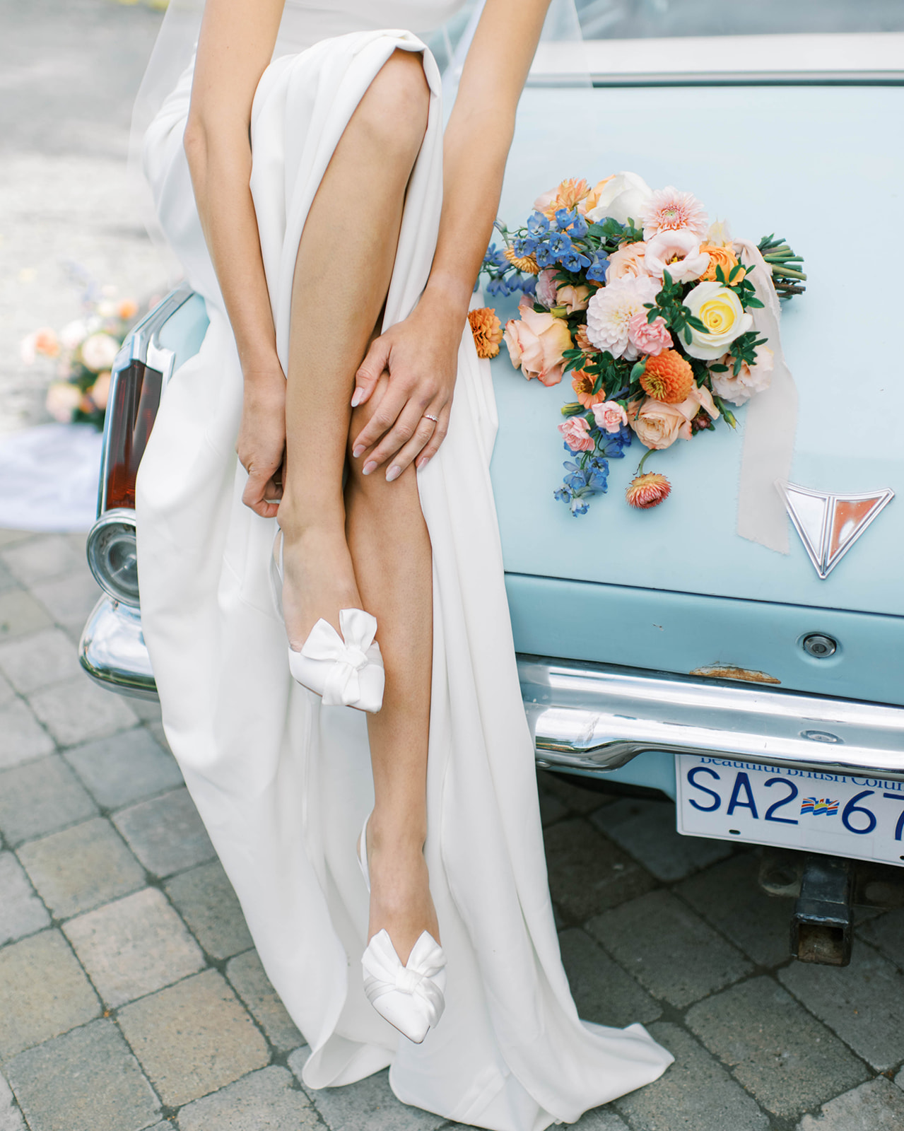 Bride adjusting white satin heels while sitting on the back of a baby blue vintage car, bridal portrait ideas for outdoor summer wedding, summer floral style