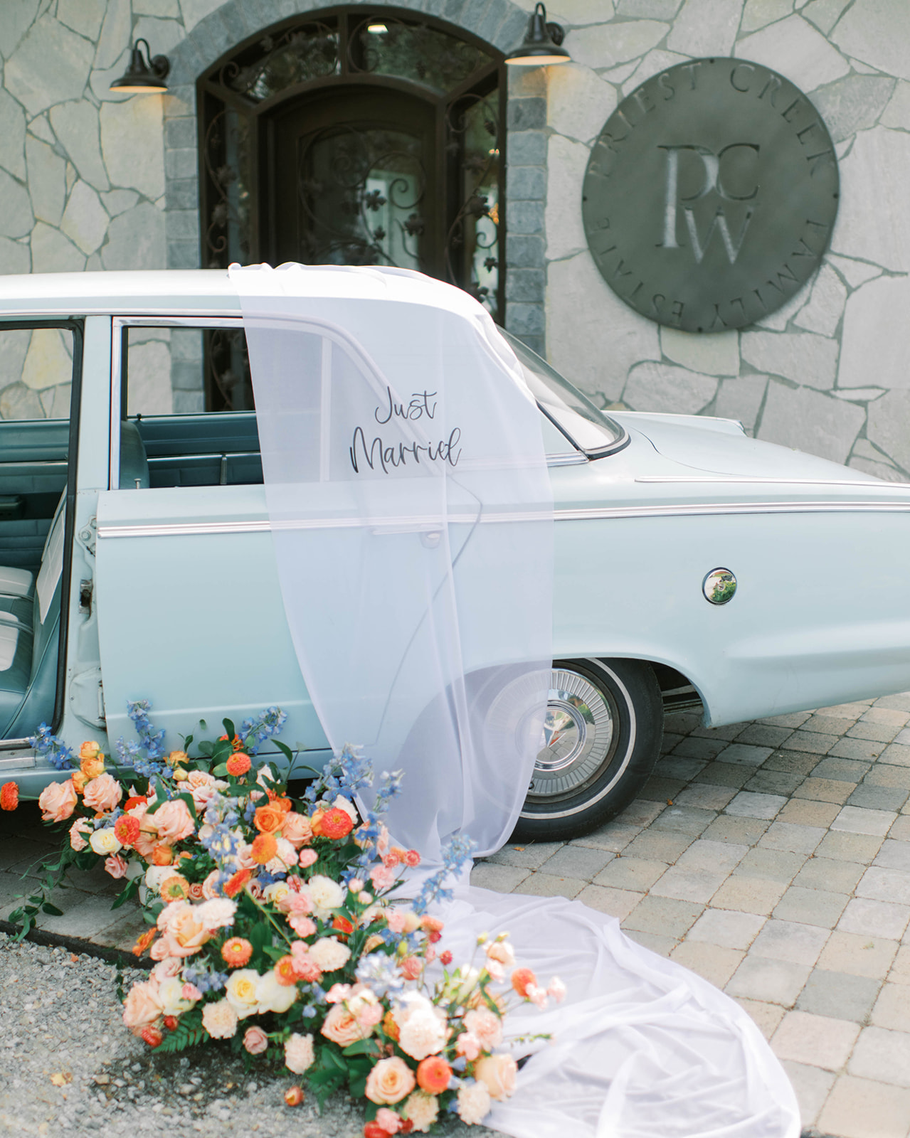 Bride's veil gracefully draped on blue vintage car with just married sign, adding a touch of romance to the getaway vehicle, lush floral arrangements adorning a classic car, transforming it into a stunning centrepiece for a vintage-inspired wedding