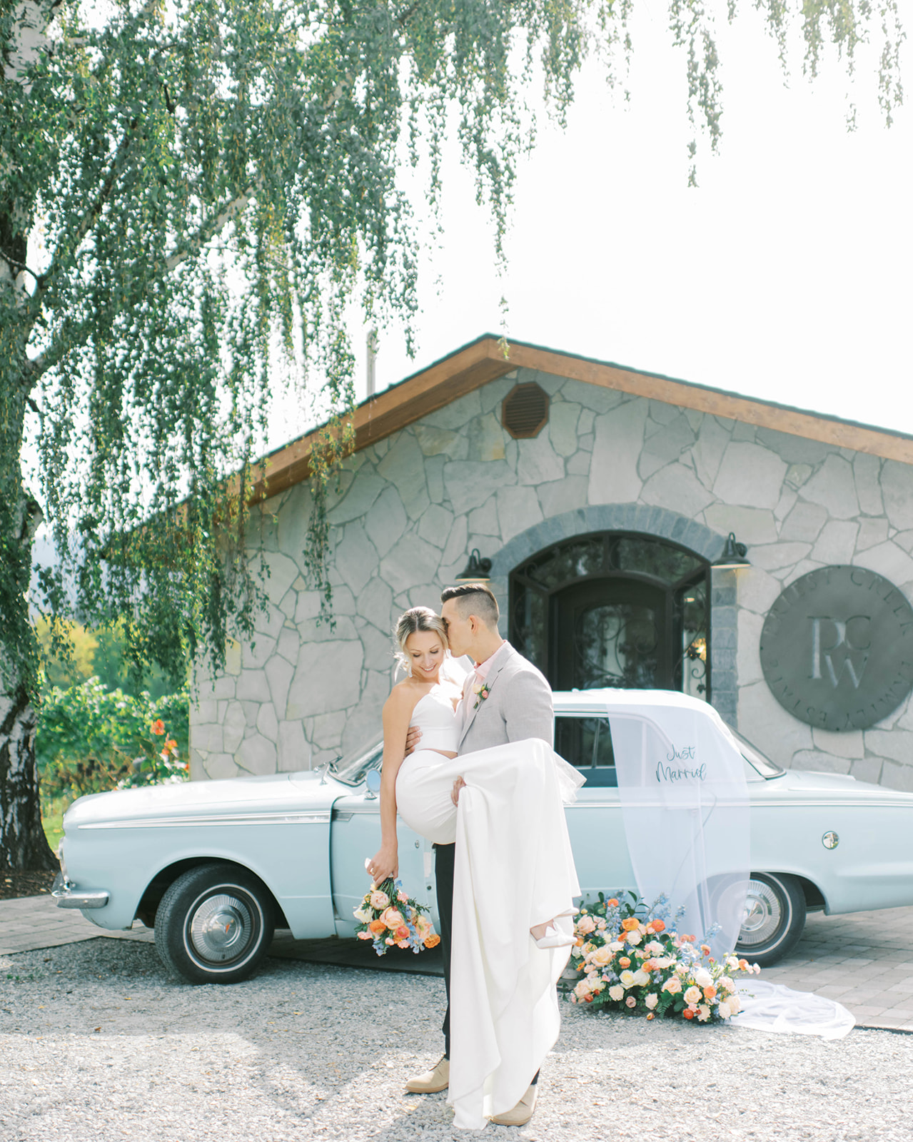 Groom carries bride in romantic couple's portrait at a Kelowna winery wedding, baby blue vintage wedding rental car